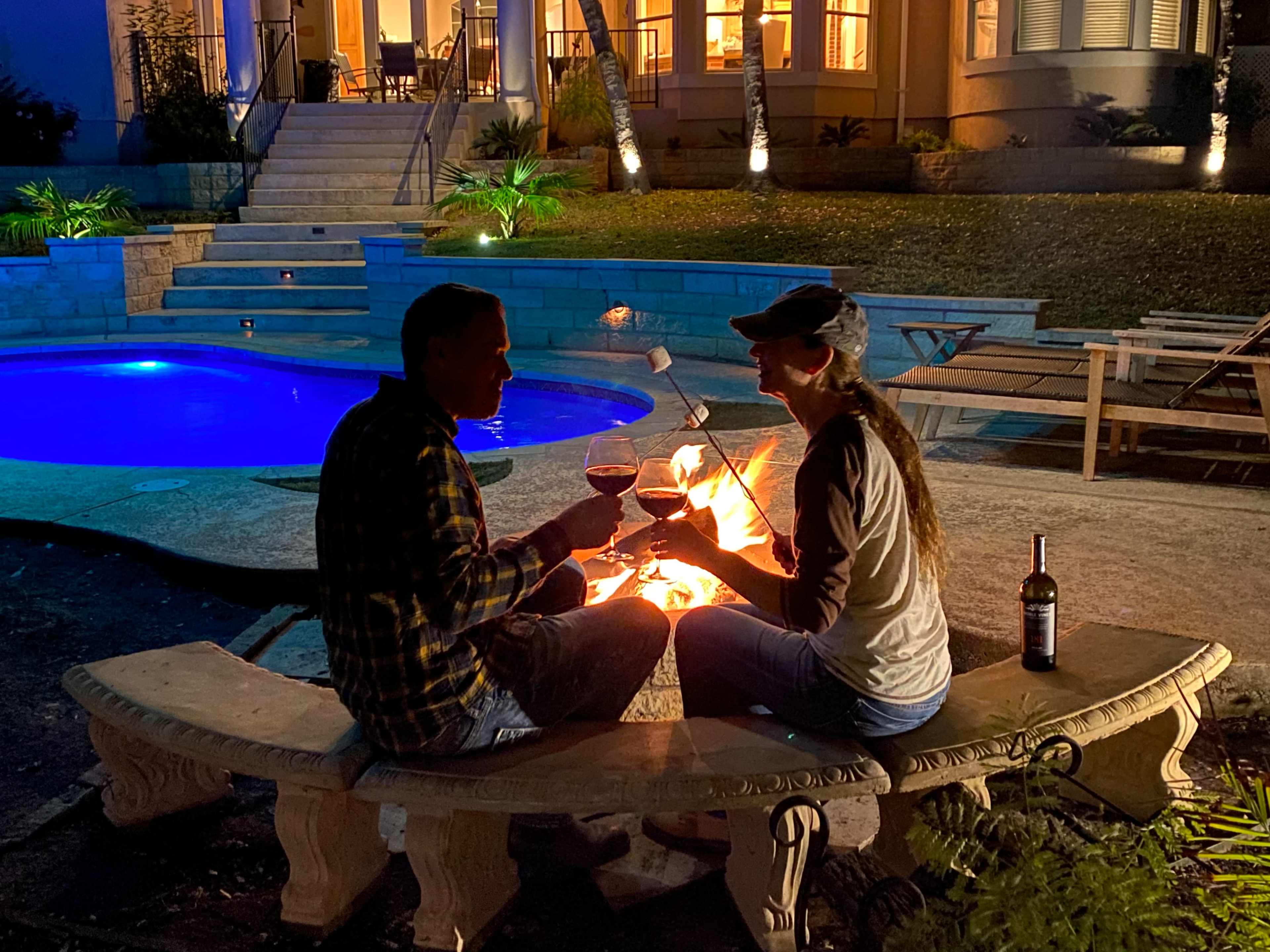 A couple toasts with glasses of wine while sitting by a fire pit in a backyard with a swimming pool and a well-lit house in the background.
