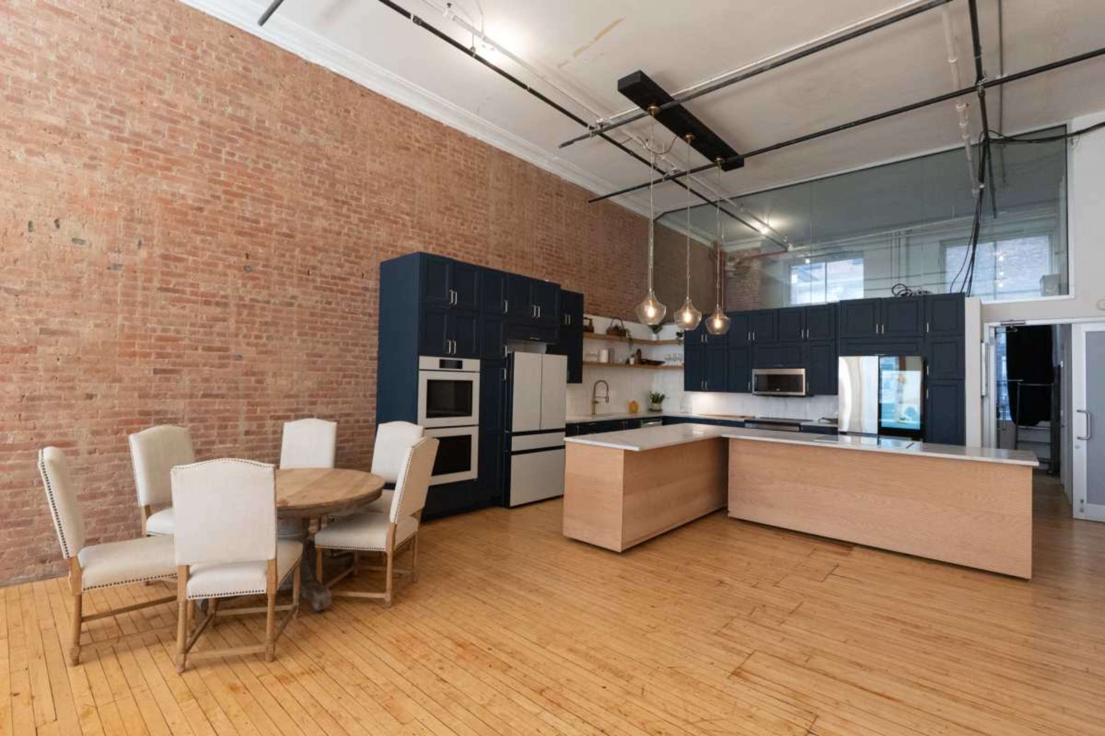 The image shows a modern kitchen with blue cabinetry, a wooden island, a circular dining table with beige chairs, and exposed brick walls.