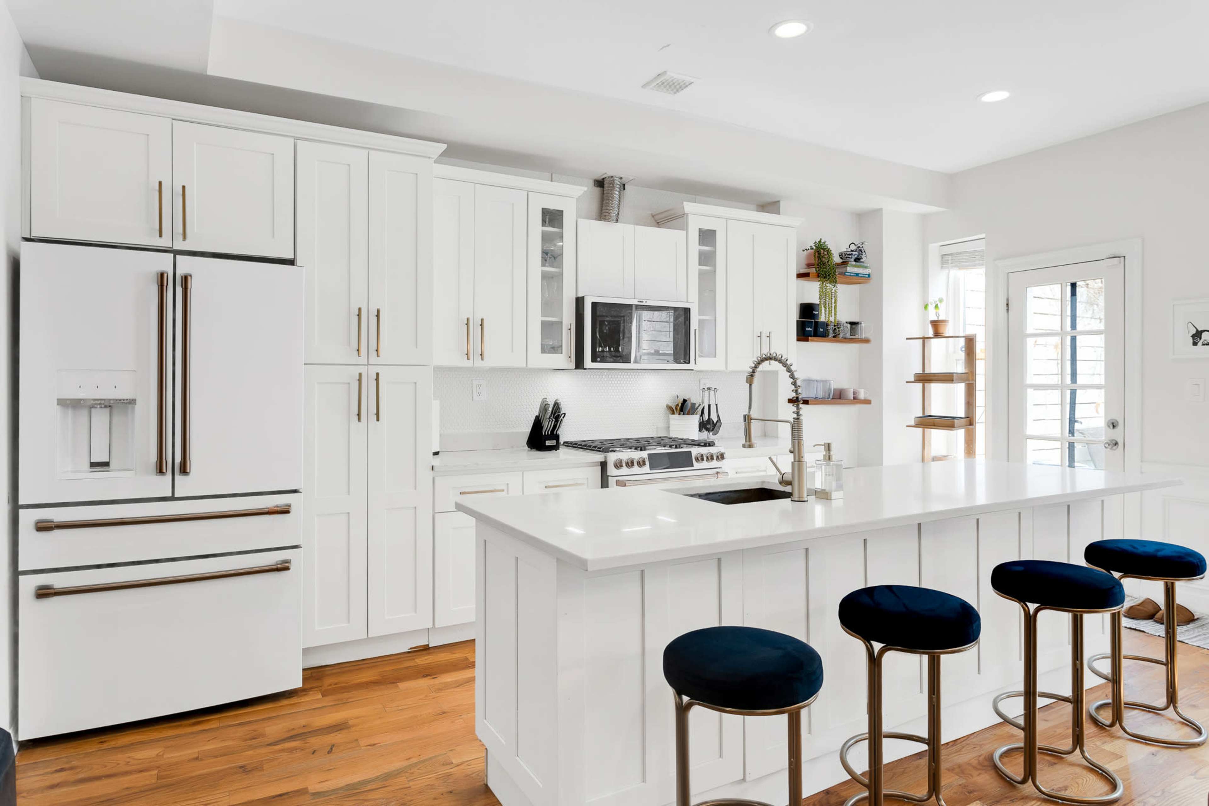 A modern kitchen featuring white cabinetry, stainless steel appliances, a central island with four blue bar stools, and a door leading outside.