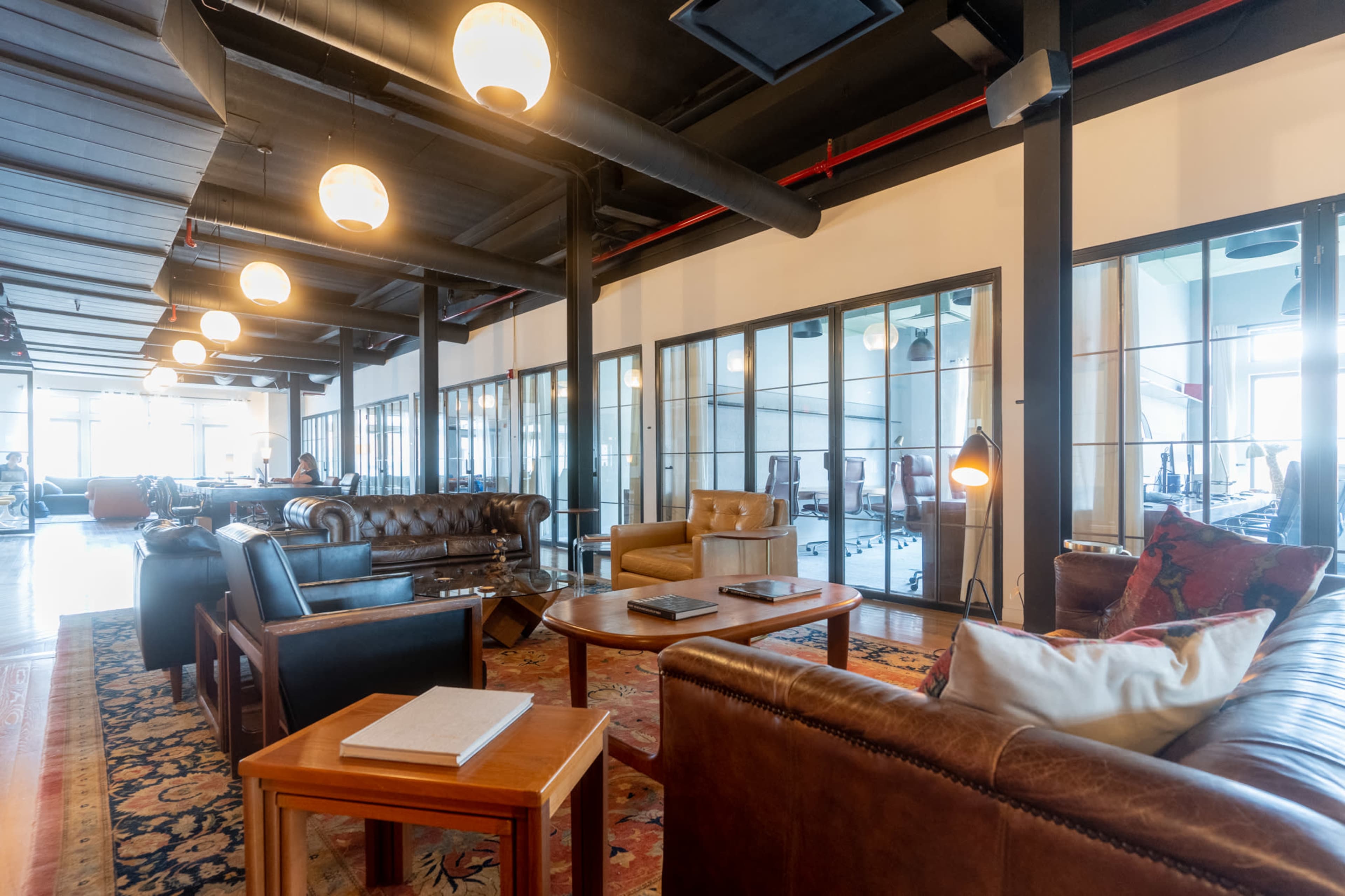 The image shows a modern office space featuring cozy seating areas with leather sofas and chairs, surrounded by glass-walled meeting rooms.