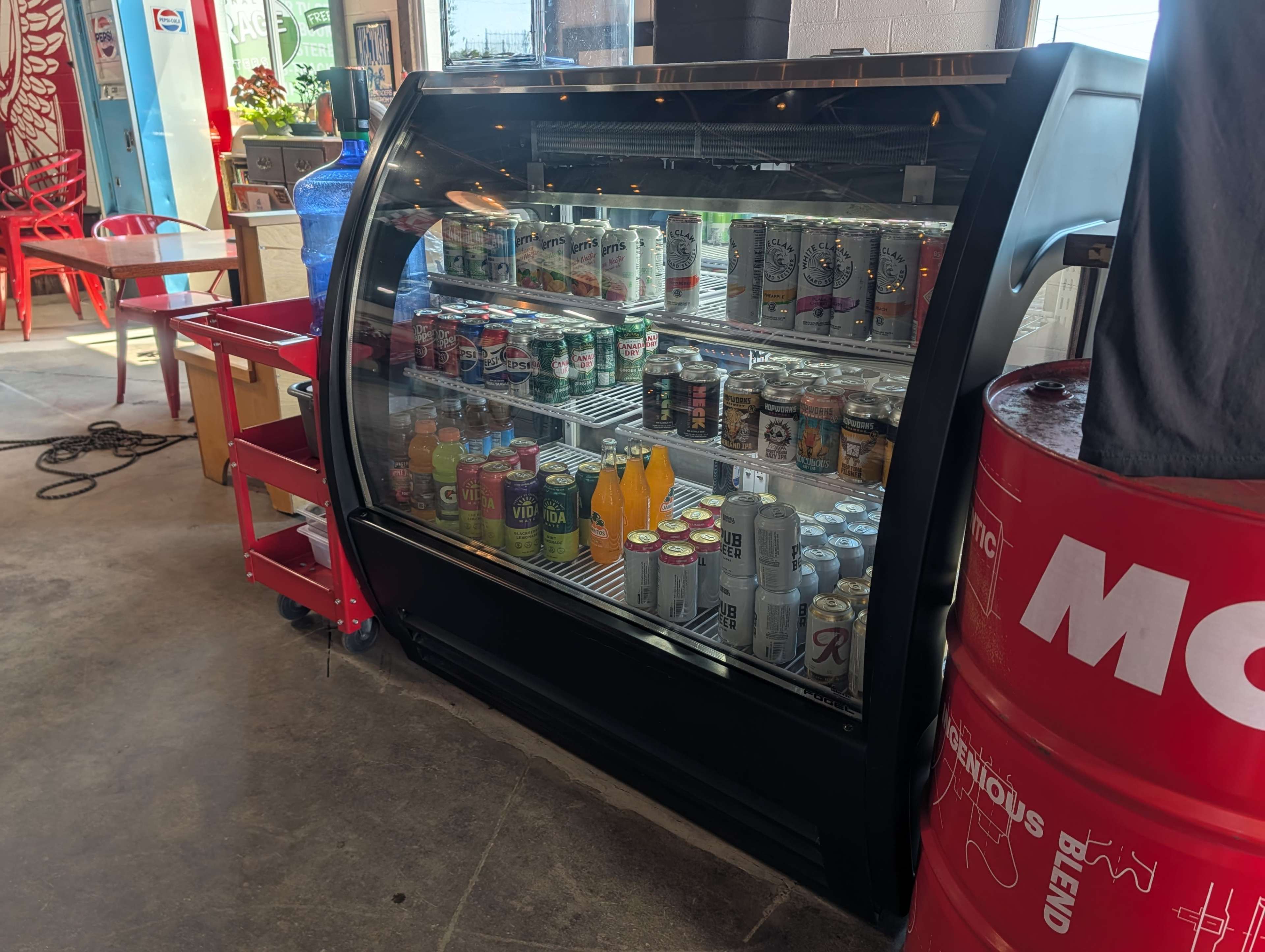 The image shows a beverage cooler filled with various cans and bottles, located in a vibrant indoor setting with red chairs and a colorful wall.
