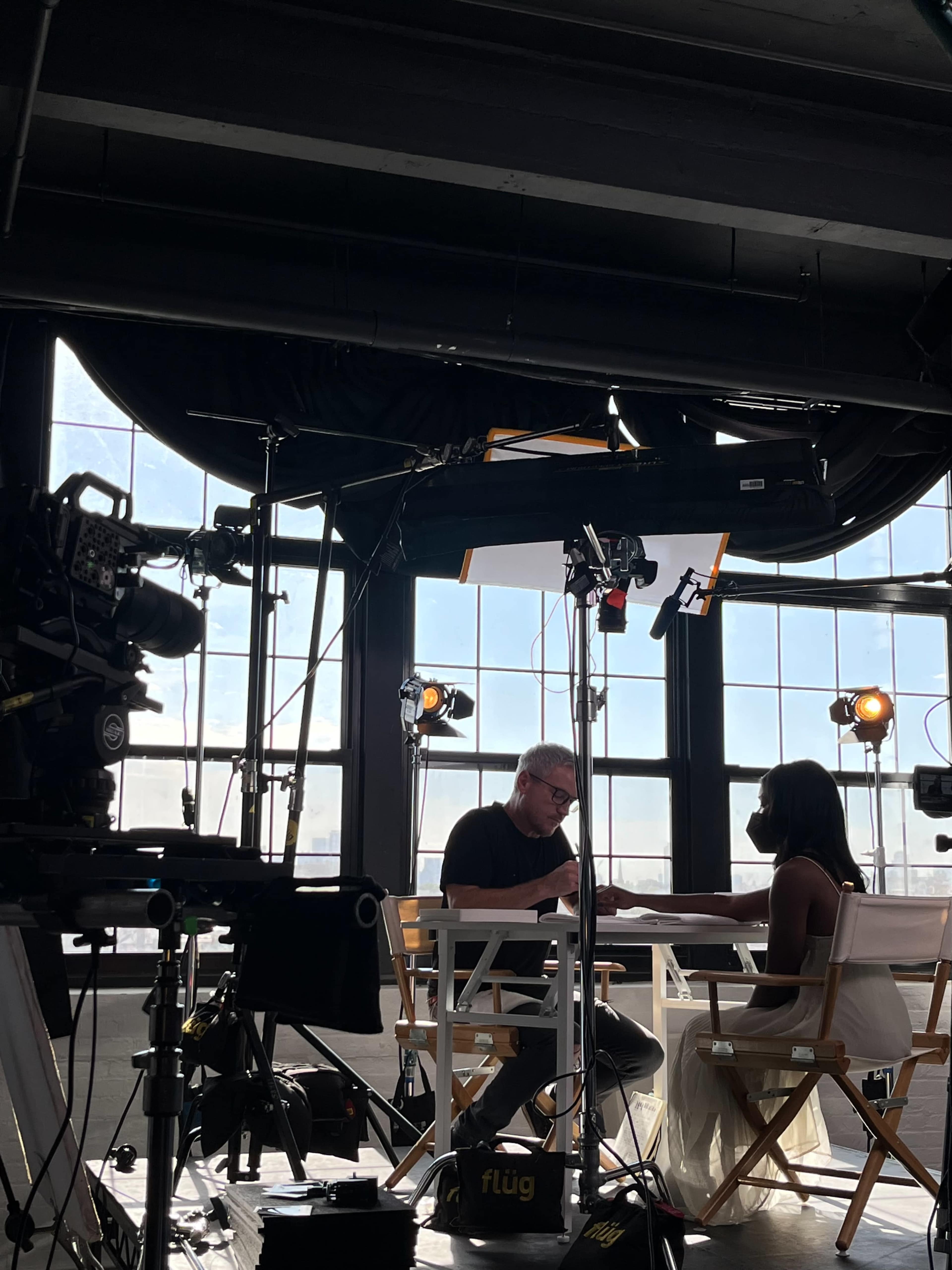 A man and woman are seated at a table with cameras and lights set up in a bright studio space.