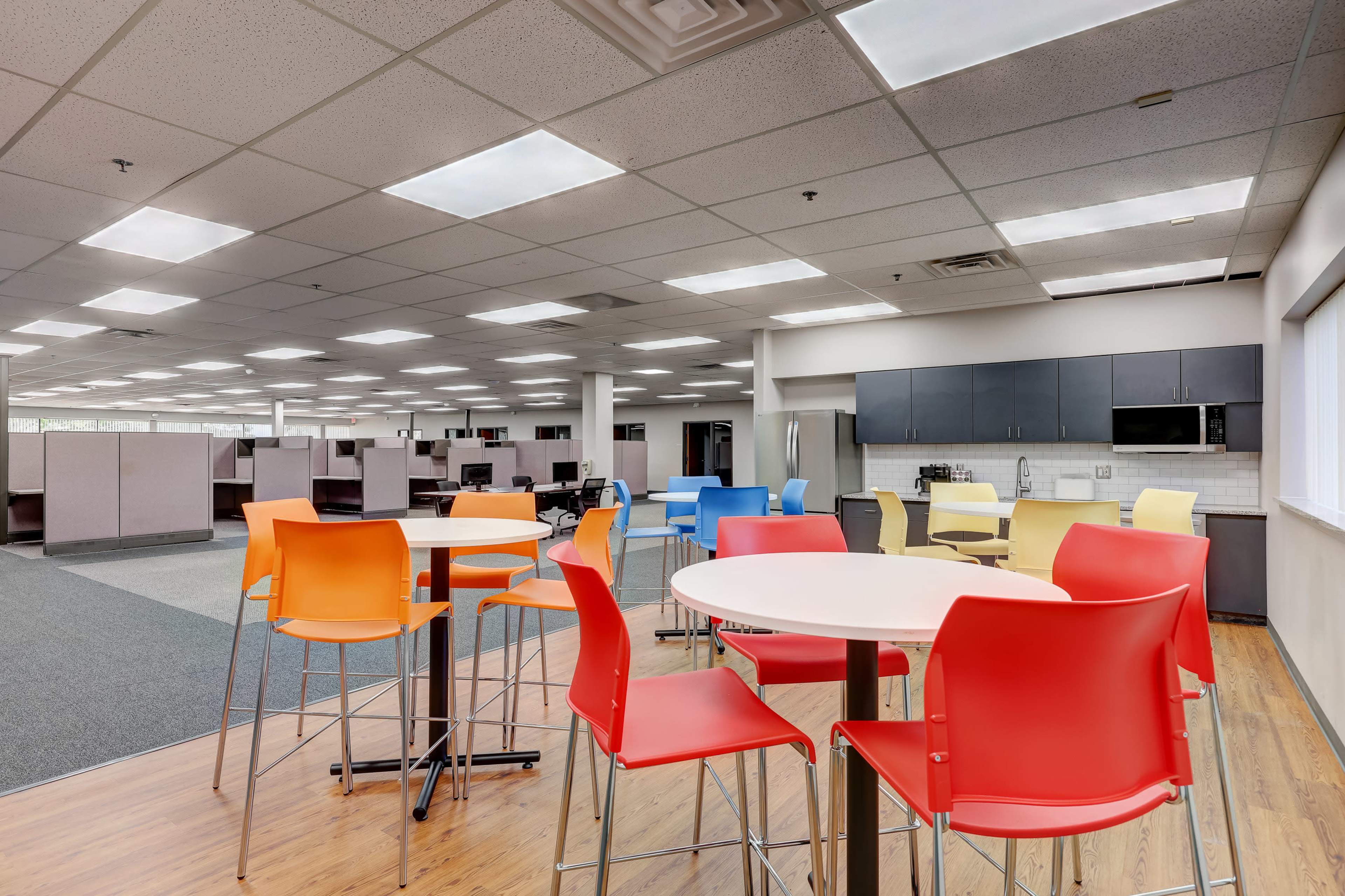 A contemporary office break area featuring colorful chairs around tables, with cubicles and workstations visible in the background.