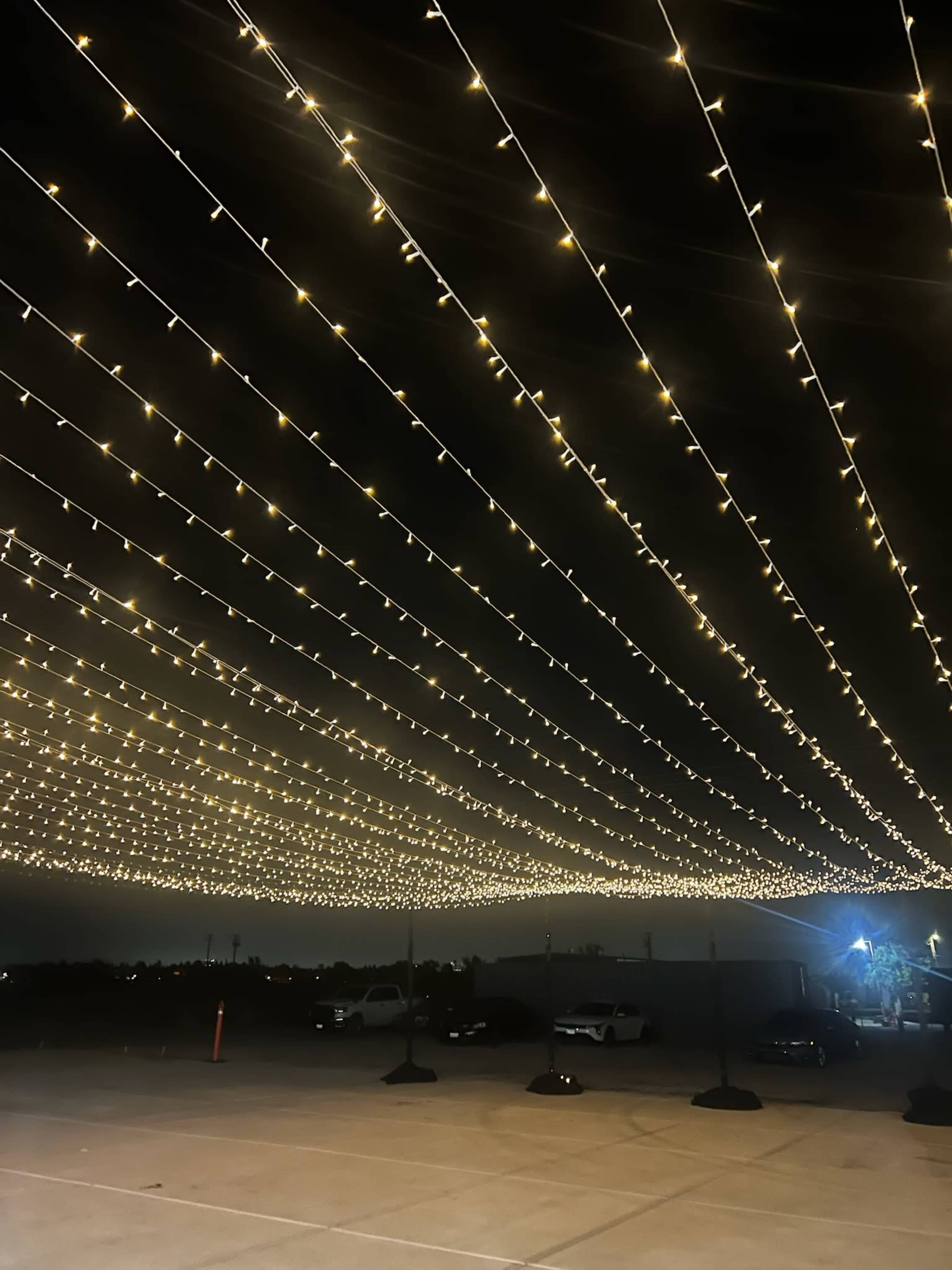 String lights are suspended overhead in a nighttime setting, illuminating a parking area beneath.