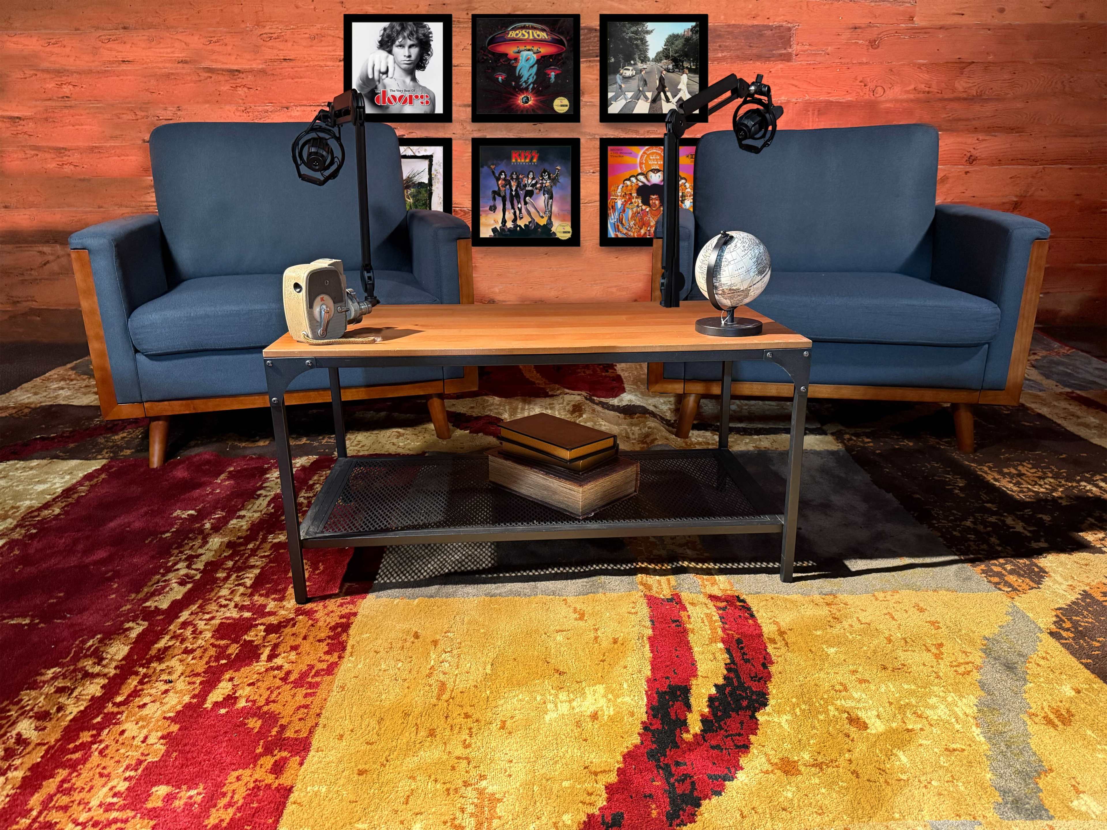 The image shows a cozy seating area with two blue armchairs flanking a wooden coffee table, positioned on a colorful rug, with a backdrop of framed albums on a wooden wall.