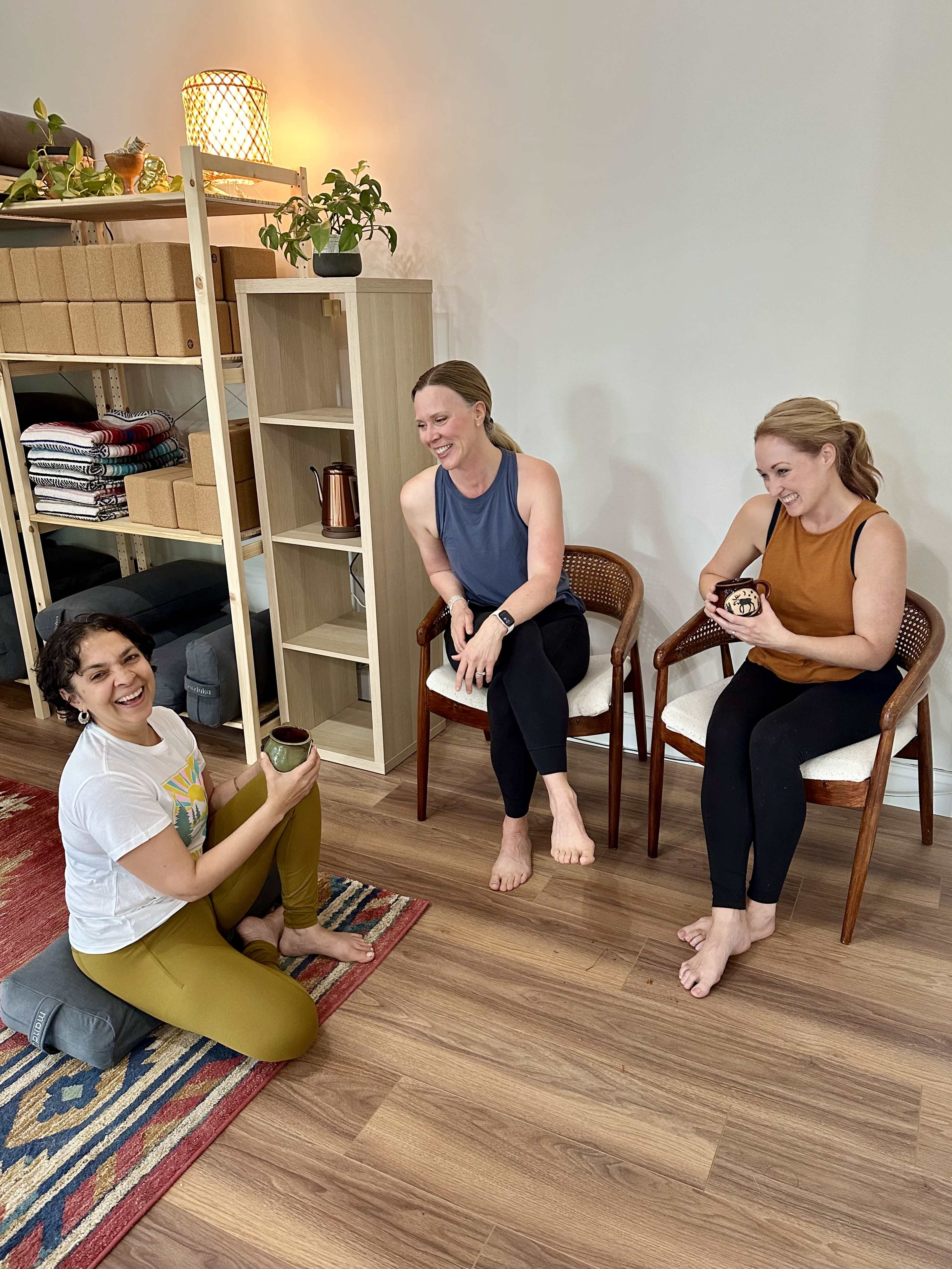Three women are seated in a room engaging in conversation while one sits on the floor and the others are on chairs.