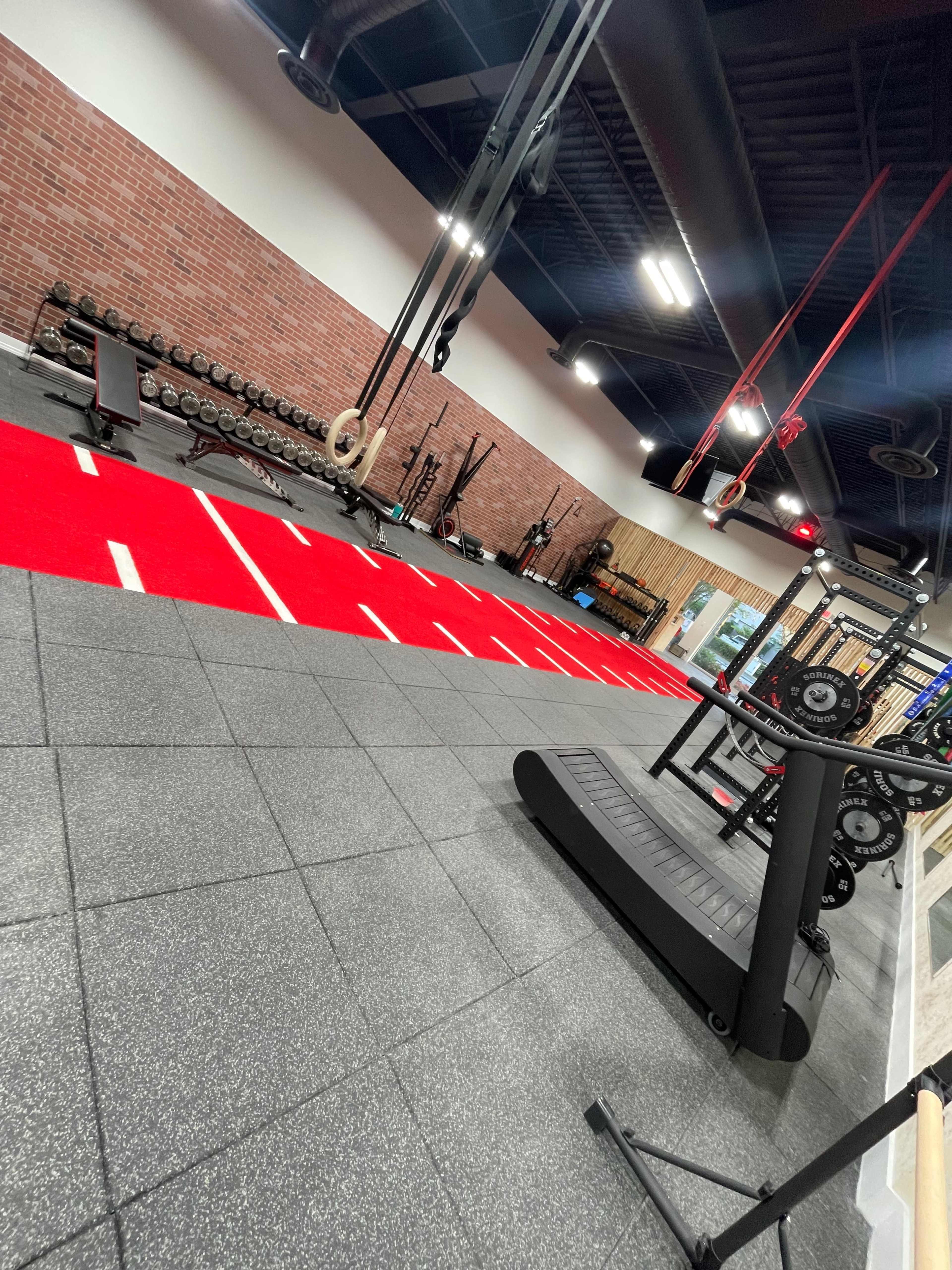 A fitness gym with weightlifting equipment, a treadmill, and a red-marked track on a rubber flooring surface.