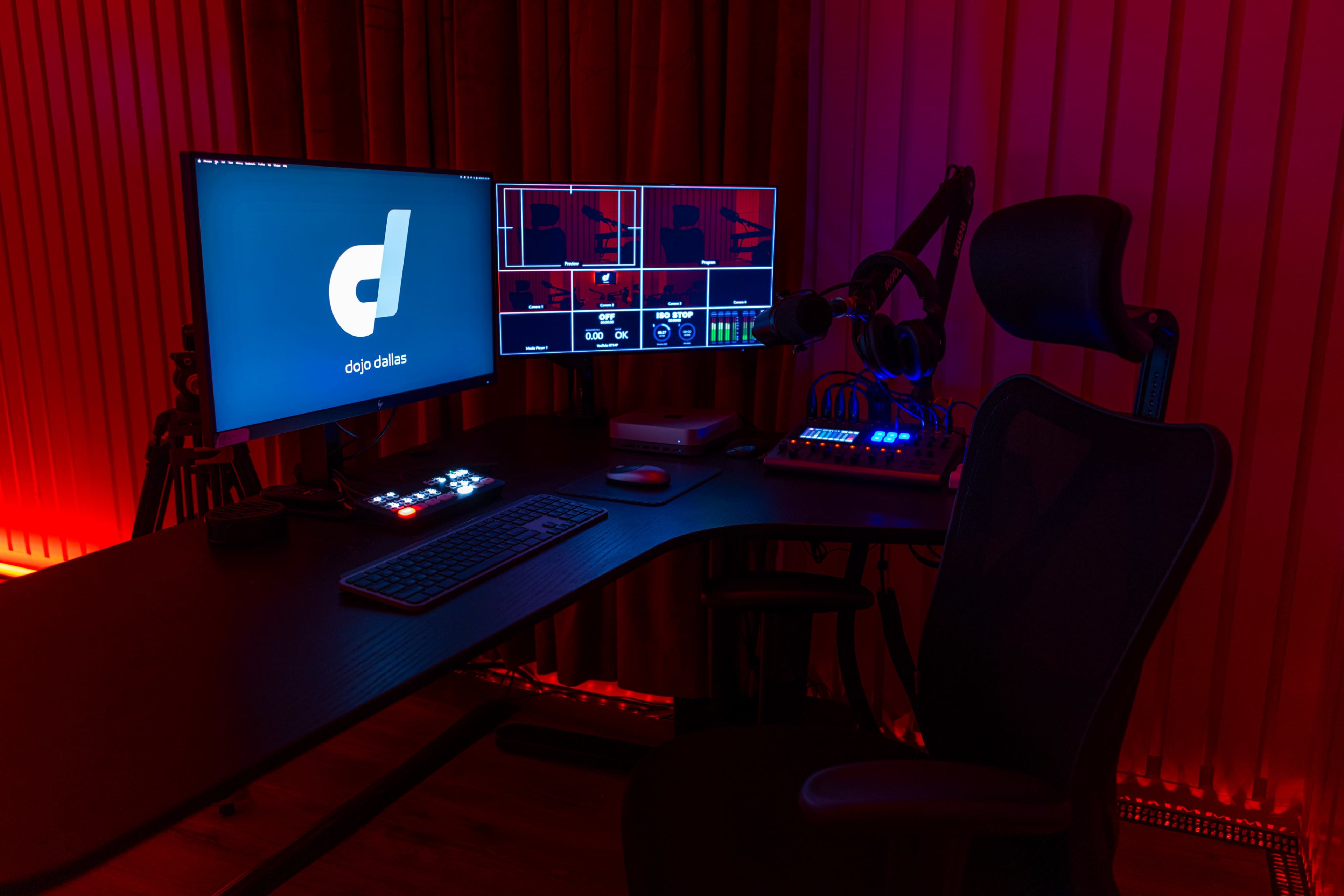 A dimly lit workspace featuring a desk with dual monitors, a keyboard, and an audio mixer, accompanied by a gaming chair and ambient red lighting.