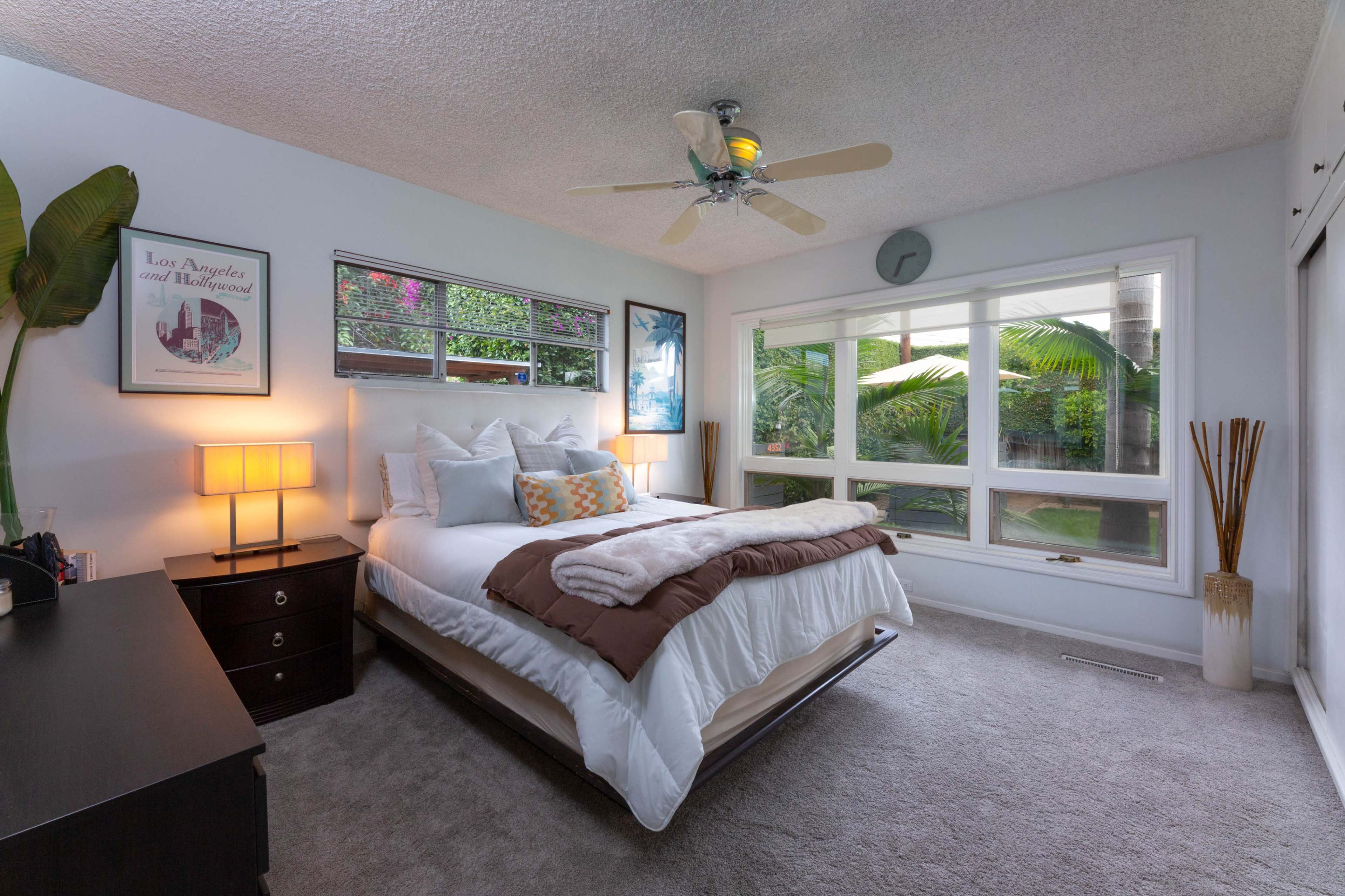 A well-decorated bedroom featuring a bed with white linens, a dresser, a ceiling fan, and large windows that provide a view of greenery outside.