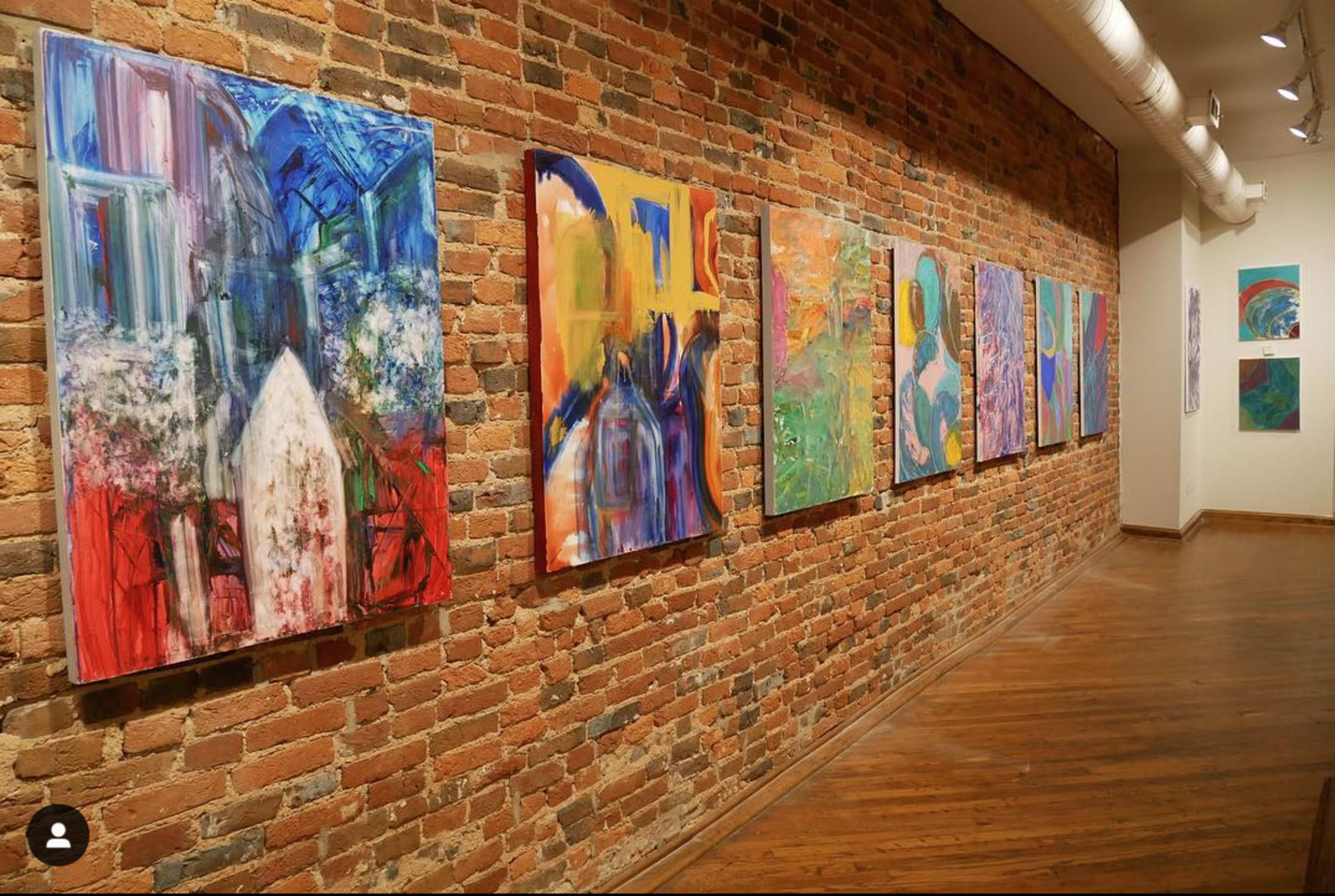 A gallery wall with a series of colorful paintings displayed alongside a brick wall and wooden floor.