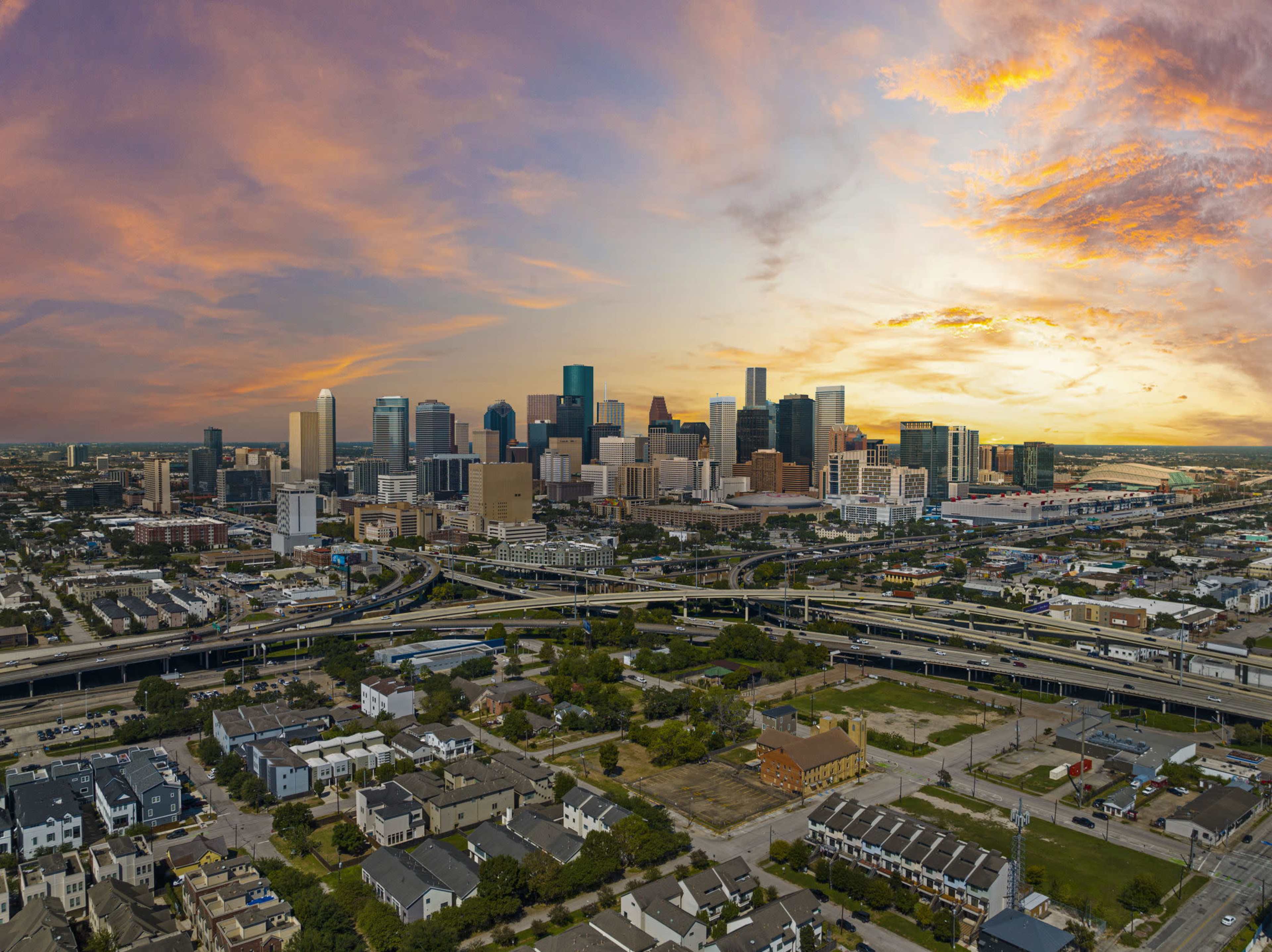 A panoramic view of a city skyline at sunset, with buildings silhouetted against a colorful sky and highways running through the foreground.