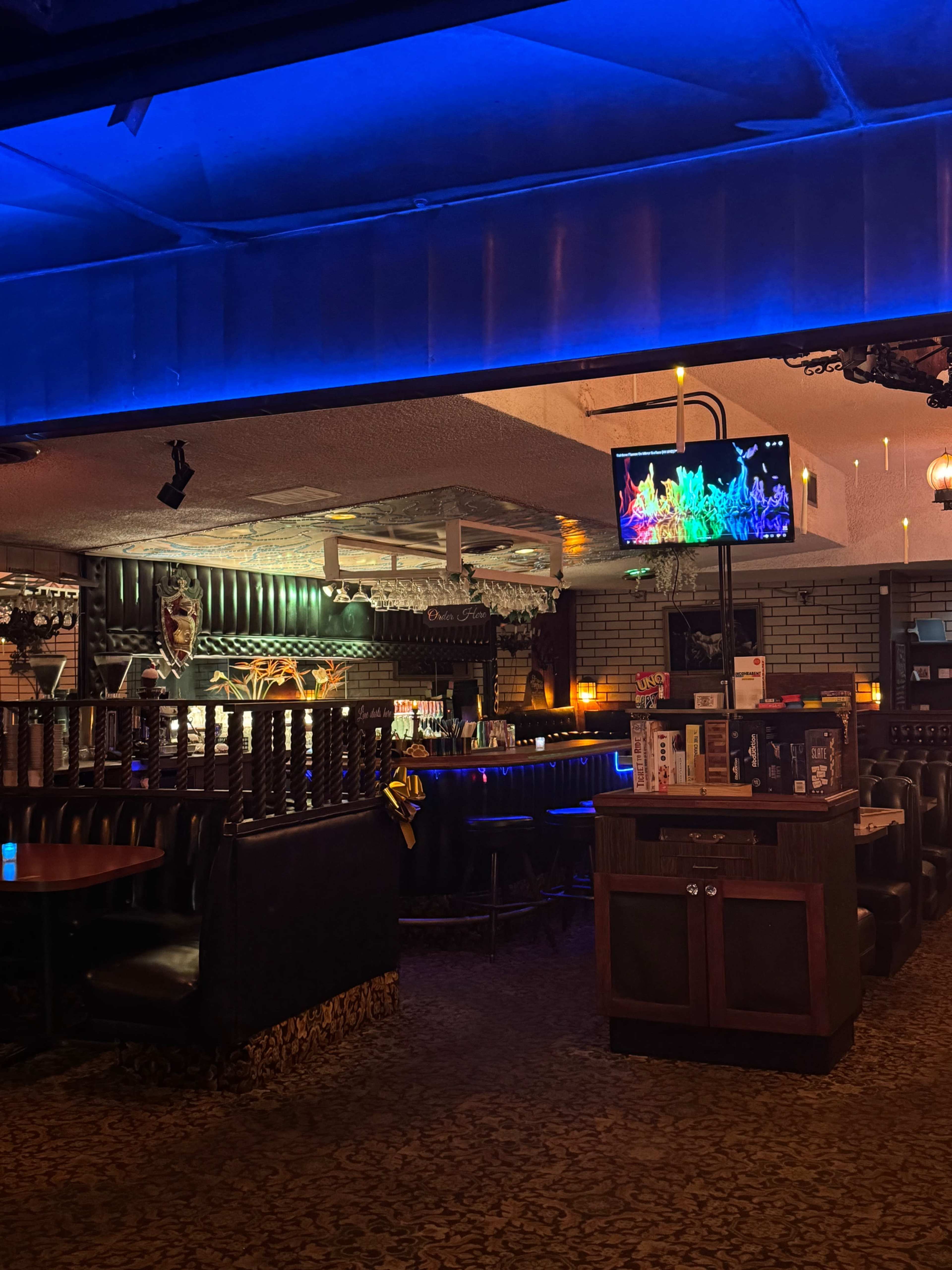 The image shows a dimly lit bar with black booths, atmospheric lighting, and a television displaying colorful visuals.