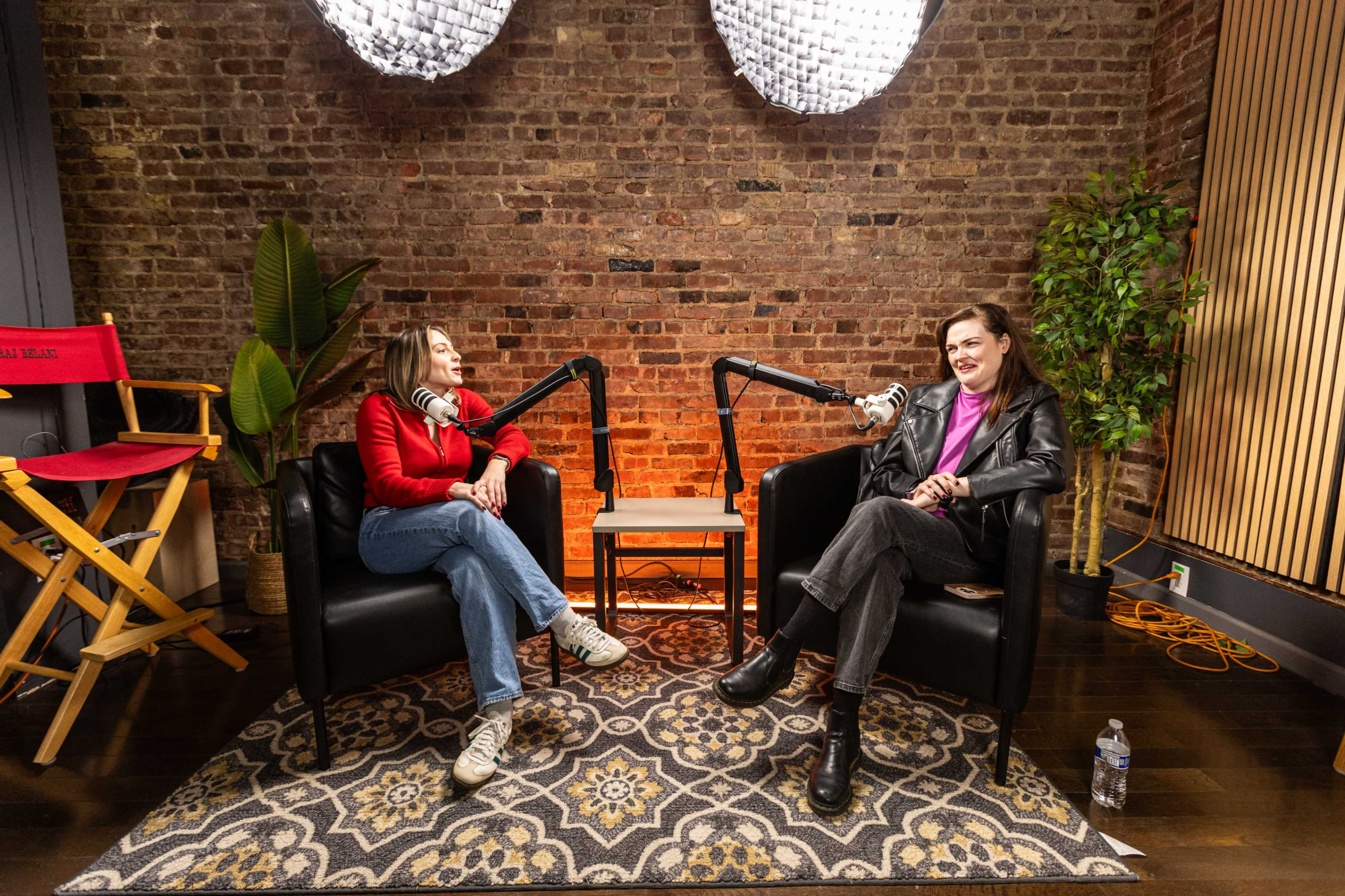 Two women sit in black chairs, engaged in conversation with microphones set up in a studio featuring a brick wall and patterned rug.