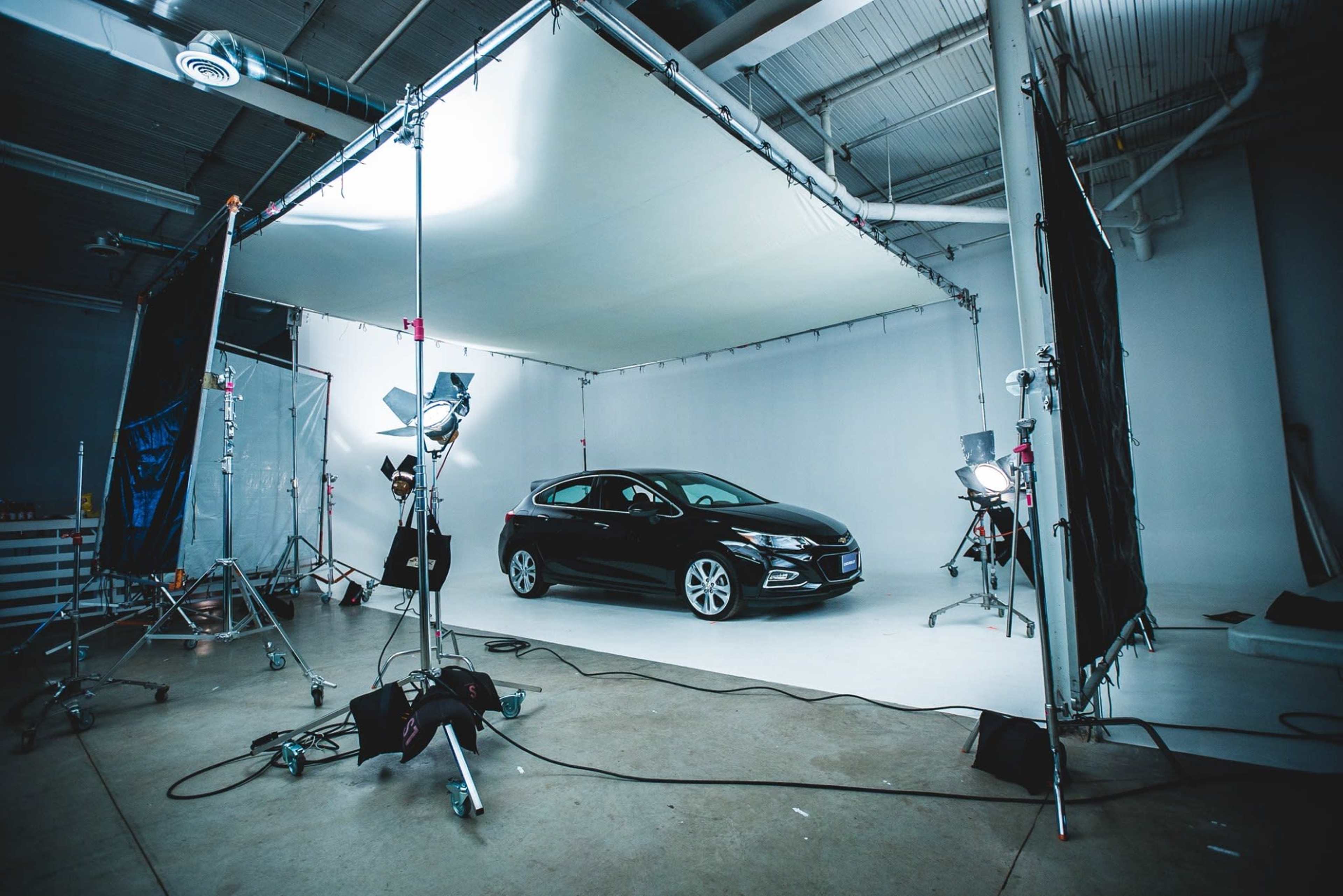 A black car is positioned in a well-lit studio on a seamless white backdrop, surrounded by photography equipment.