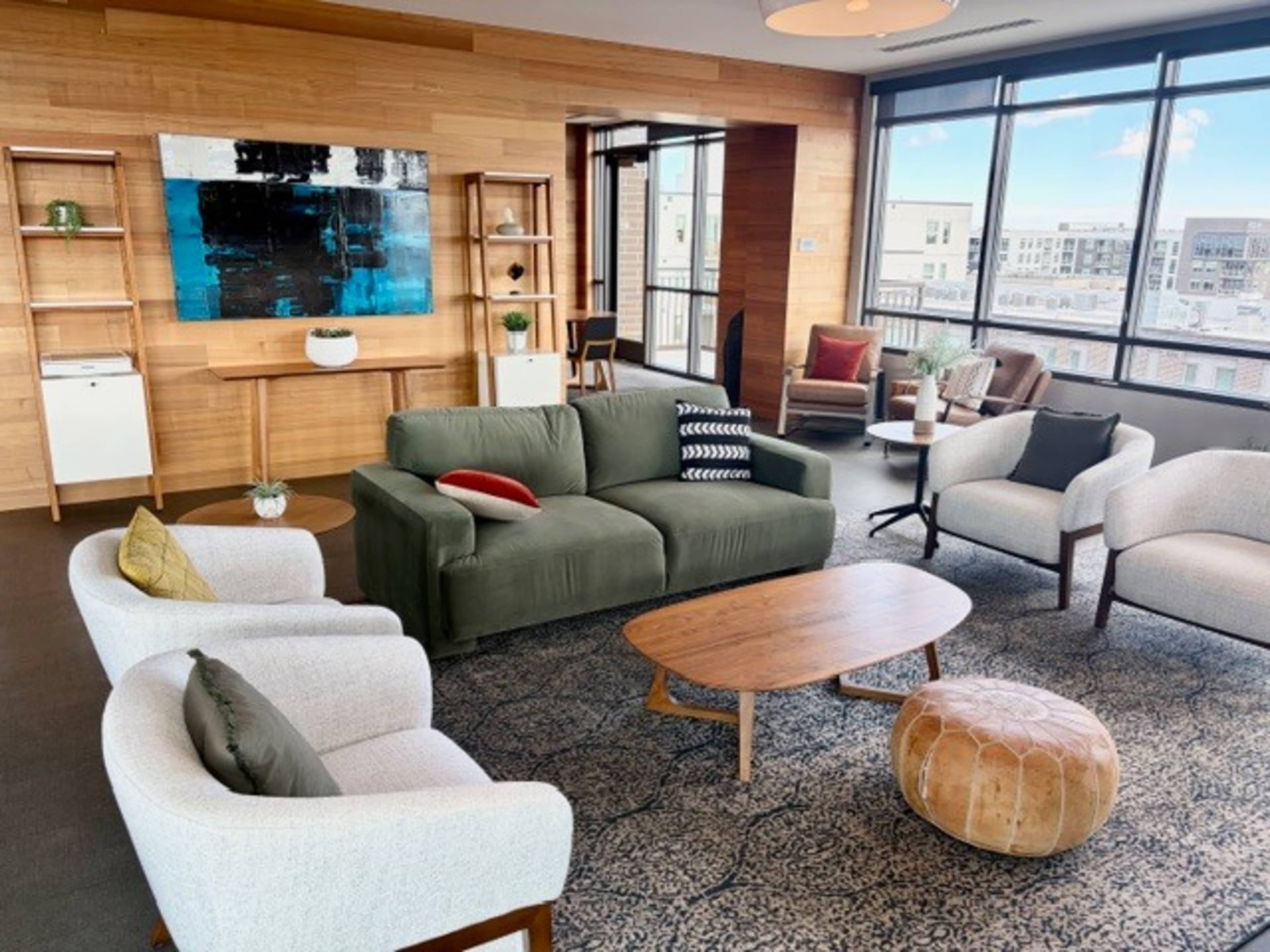 The image shows a modern living area with a green sofa, light-colored chairs, a wooden coffee table, and large windows offering a view of the city.