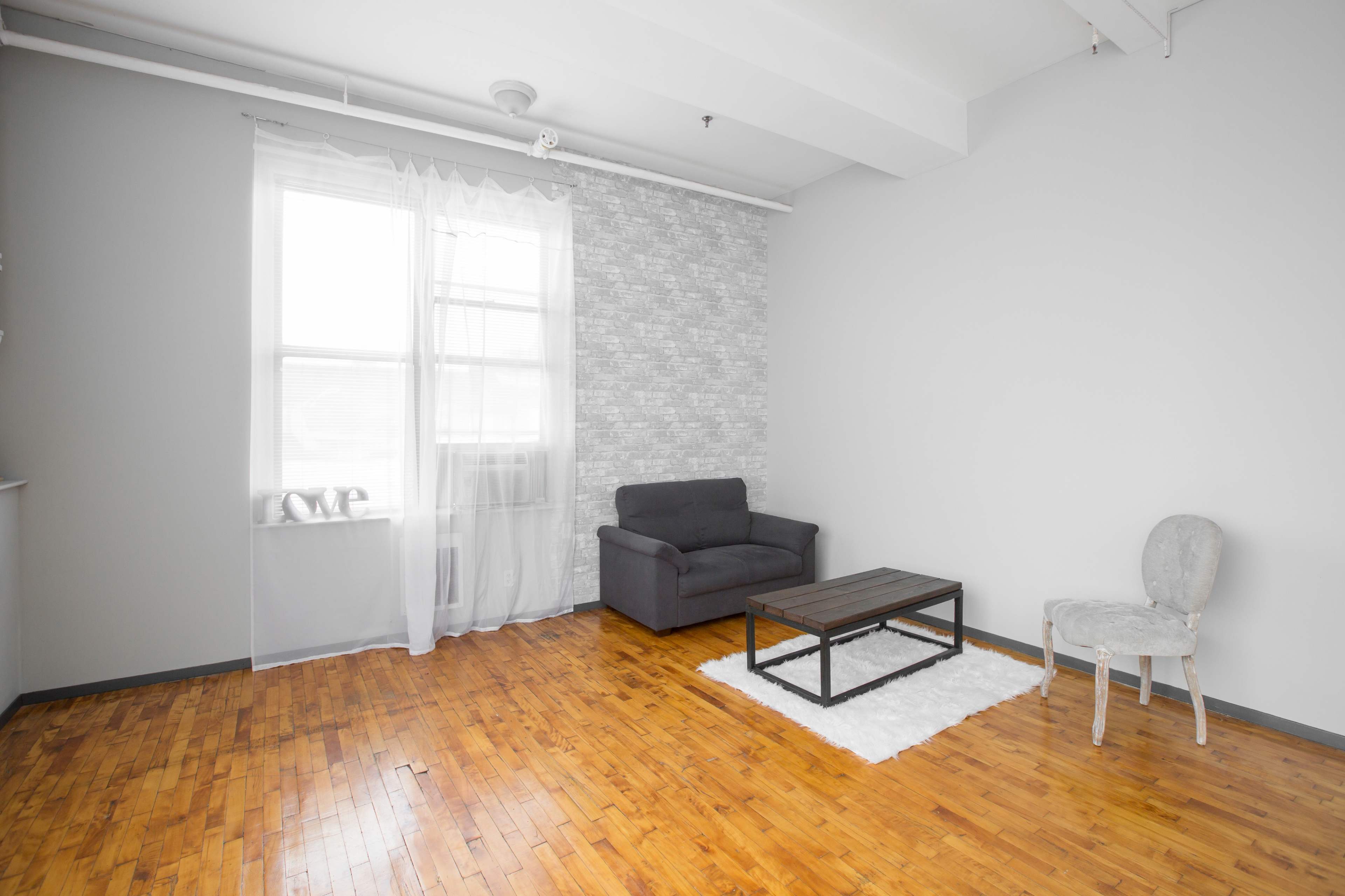 A minimalist living room features a gray chair, a wooden coffee table on a white rug, and a large window with sheer curtains.