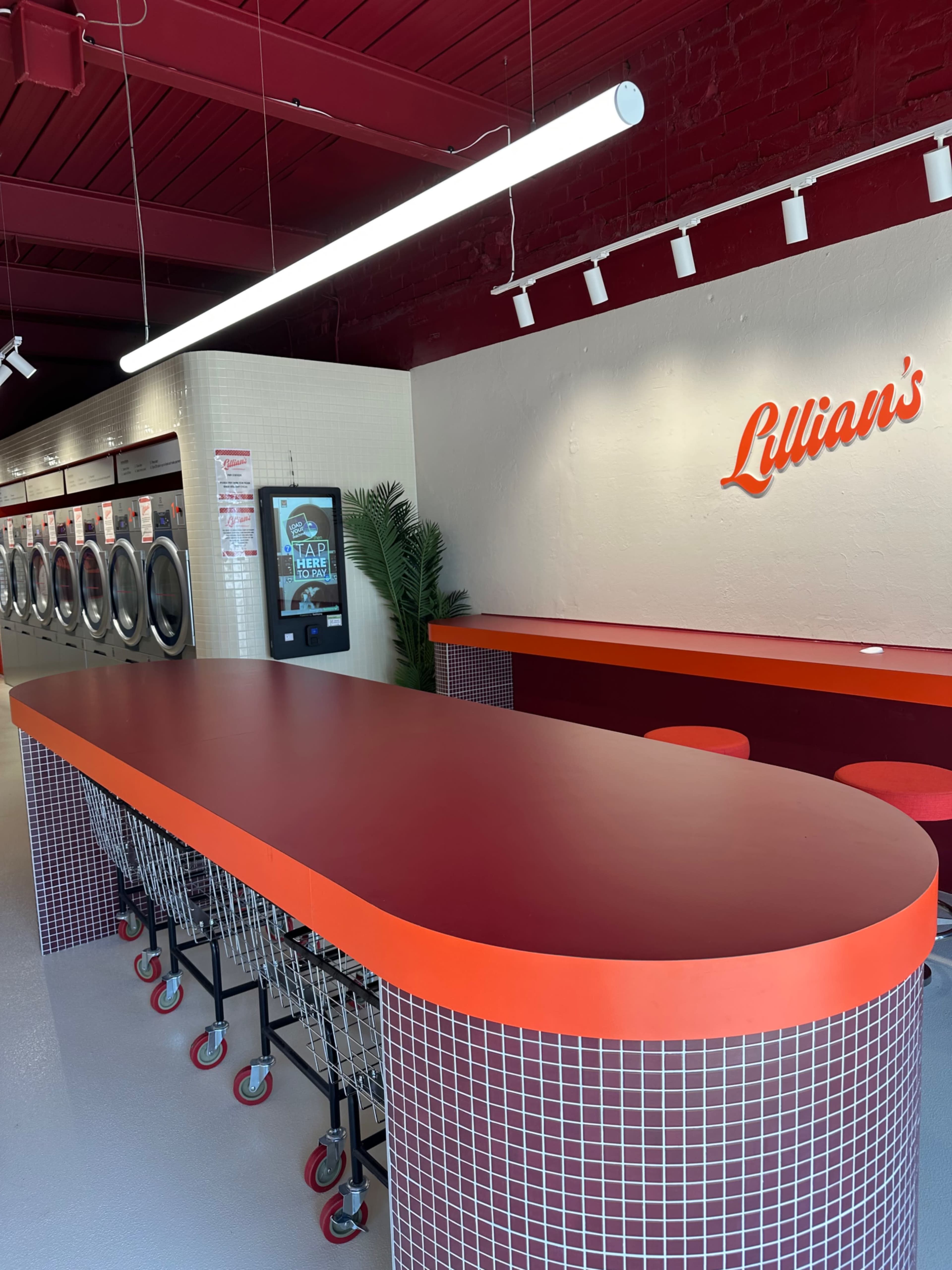 The image shows a modern laundry facility featuring a long red countertop with seating, vending machines, and washing machines along the wall.