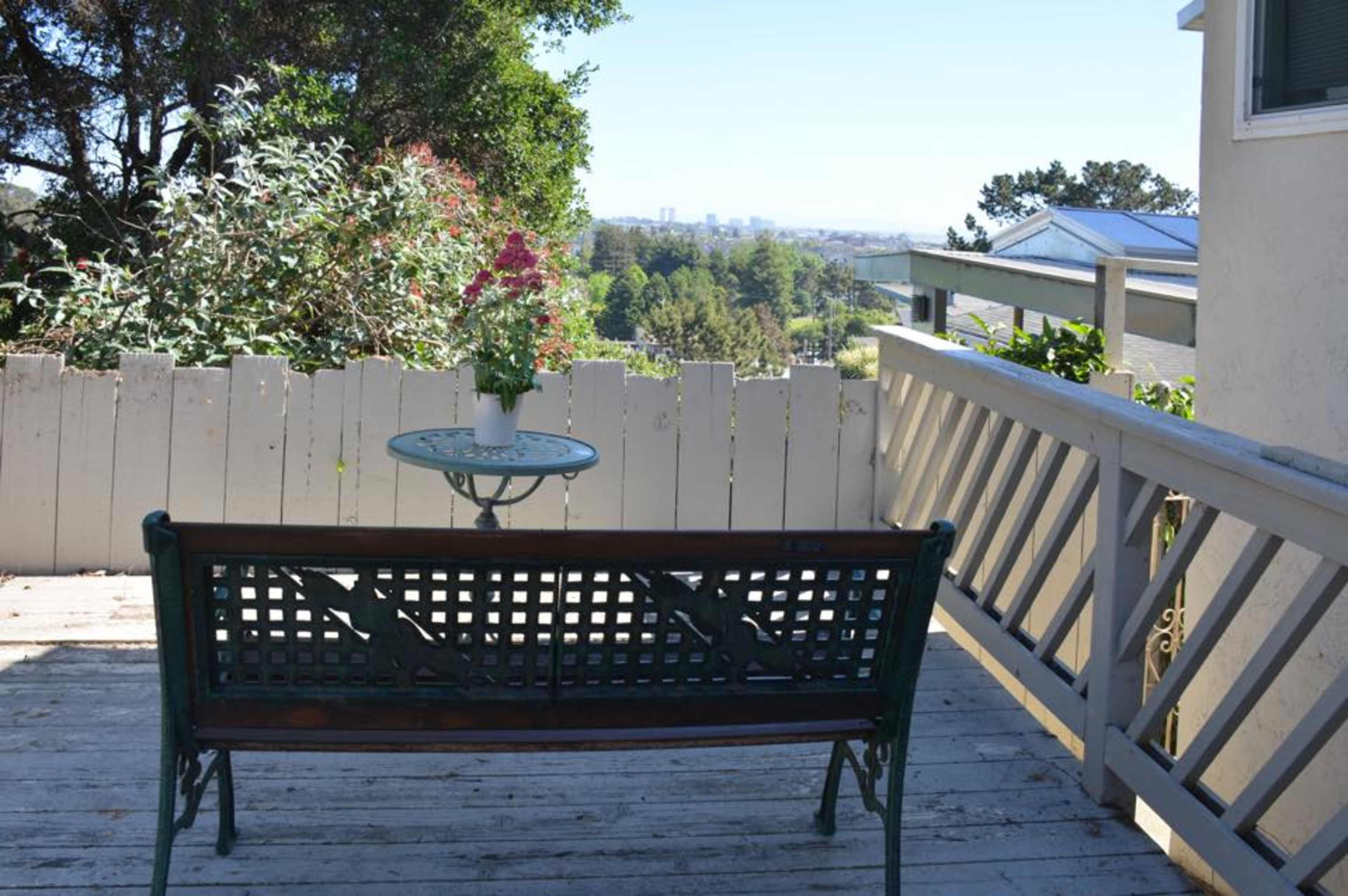 A green bench and a small table with a potted plant are positioned on a wooden deck overlooking a distant city skyline.