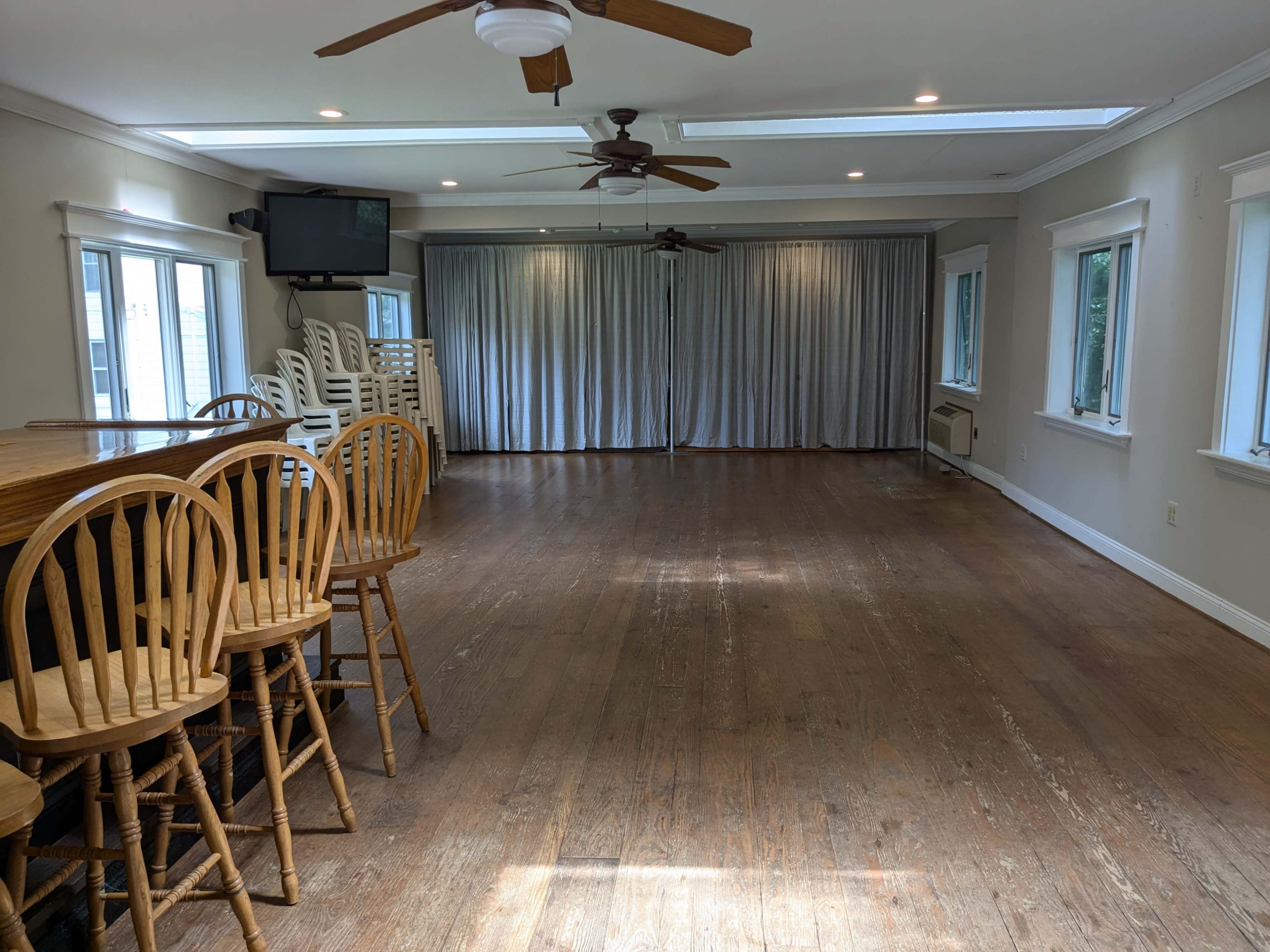 A spacious room with hardwood flooring, a bar area with wooden stools, and large curtains covering a wall of windows.