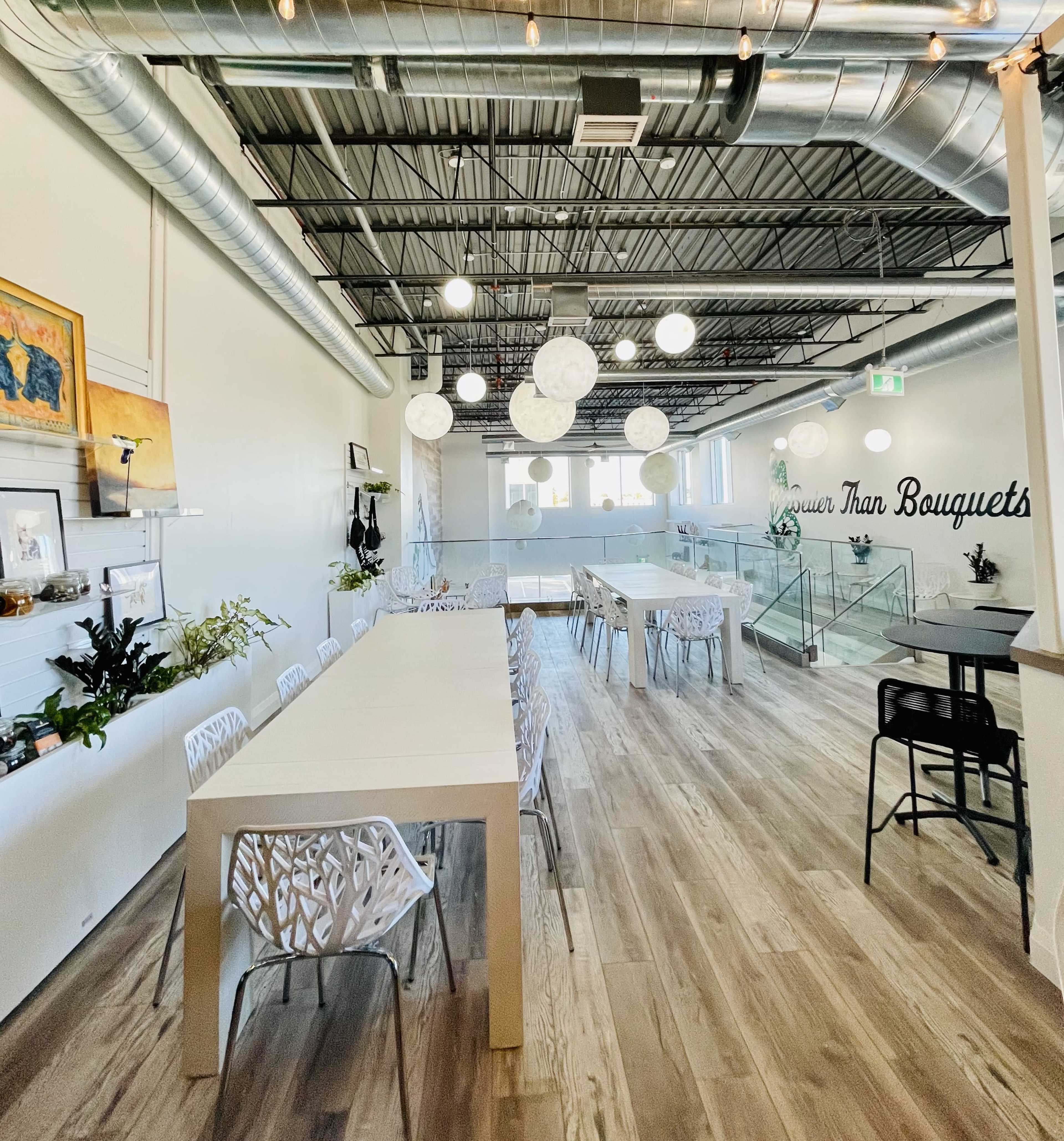 The image shows a modern, airy interior of a floral shop with long white tables, decorative pendant lights, and greenery along the walls.