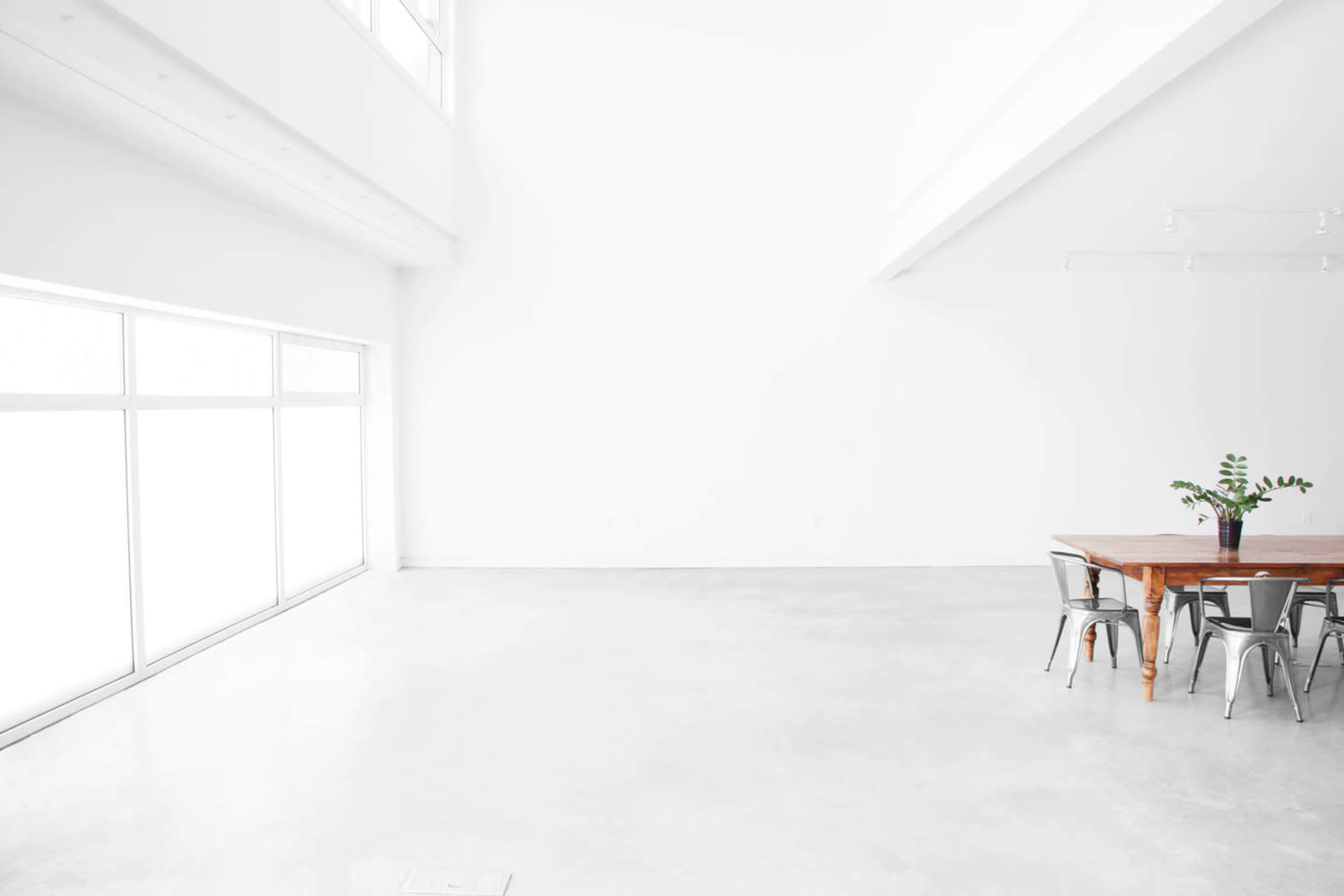 A spacious room with white walls and a polished concrete floor features a wooden table surrounded by metal chairs and large windows letting in natural light.