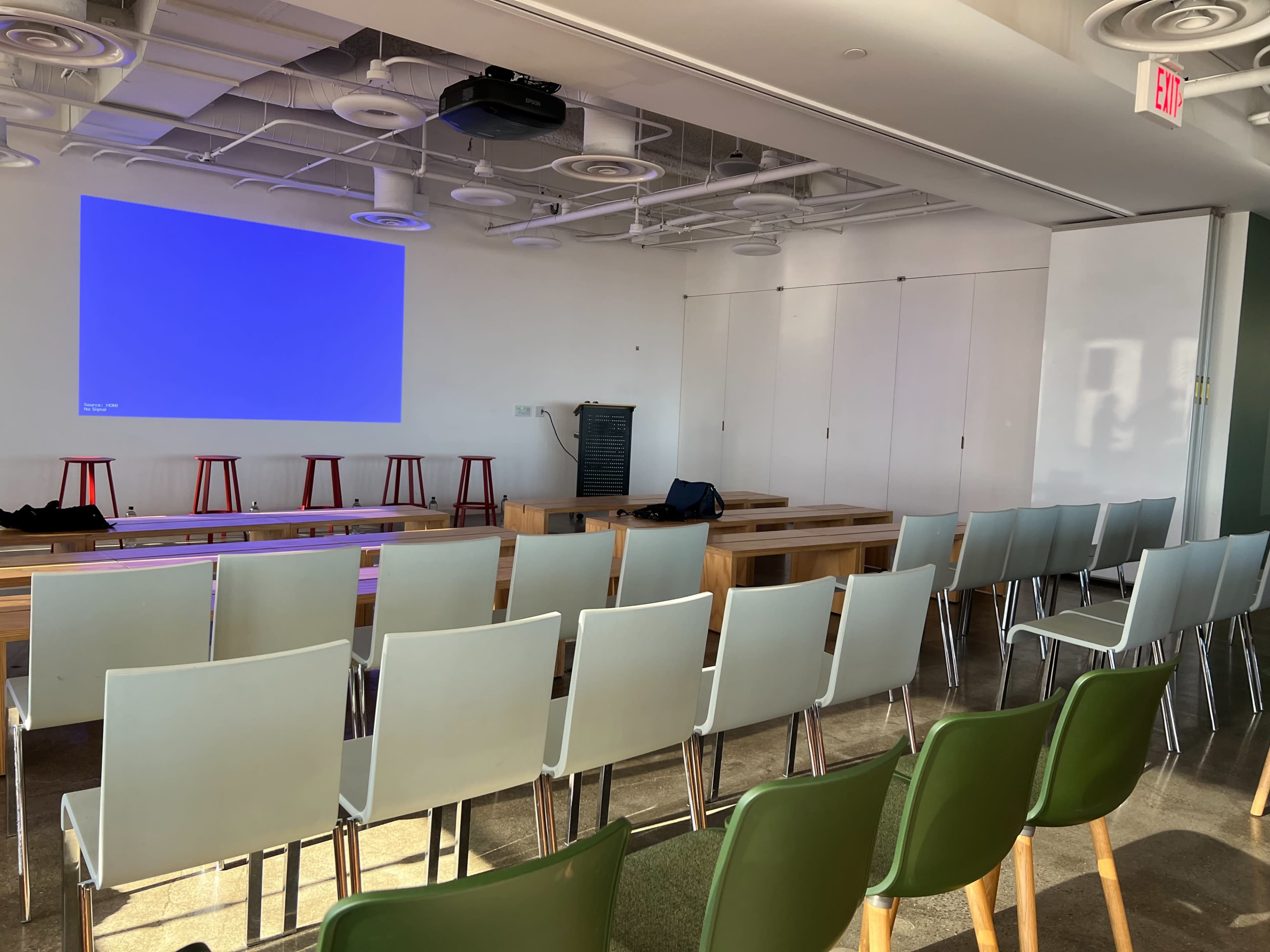A brightly lit meeting room with several rows of chairs facing a large screen at the front.