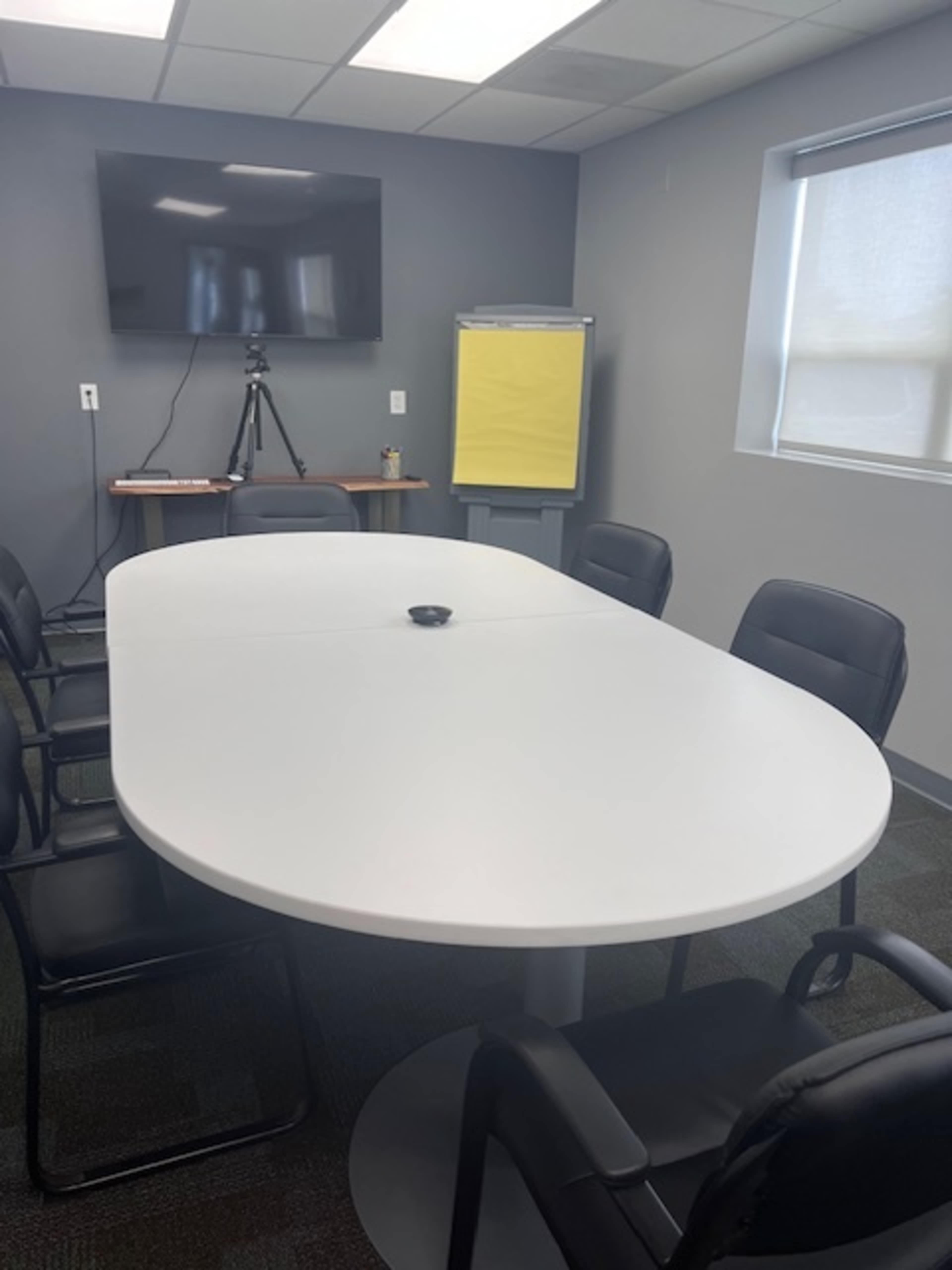 A modern conference room with a large oval table surrounded by black chairs, a wall-mounted TV, and a flip chart on an easel.