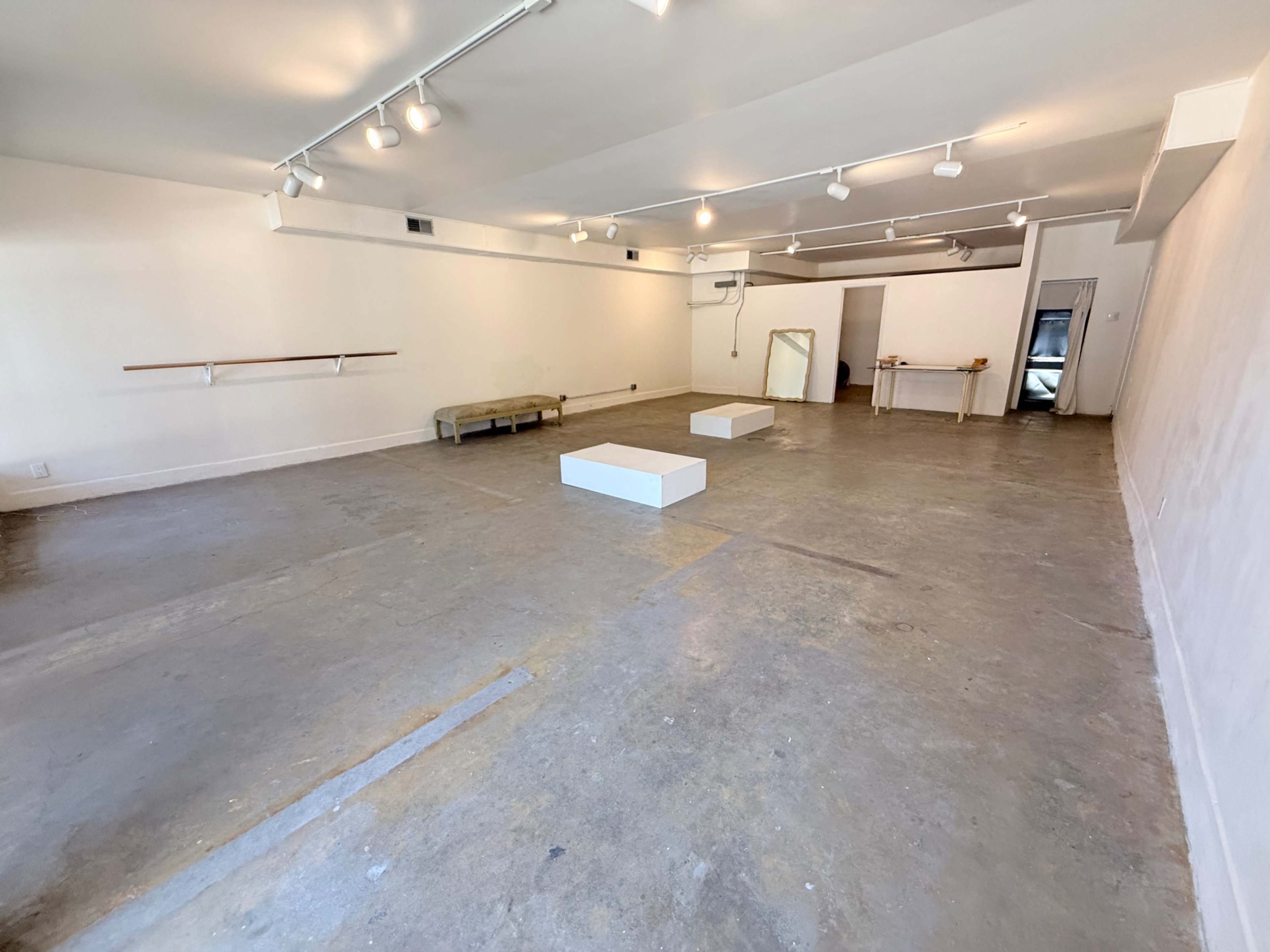 The image shows a vacant, well-lit gallery space with a concrete floor, two white pedestals in the center, and minimal furnishings along the walls.