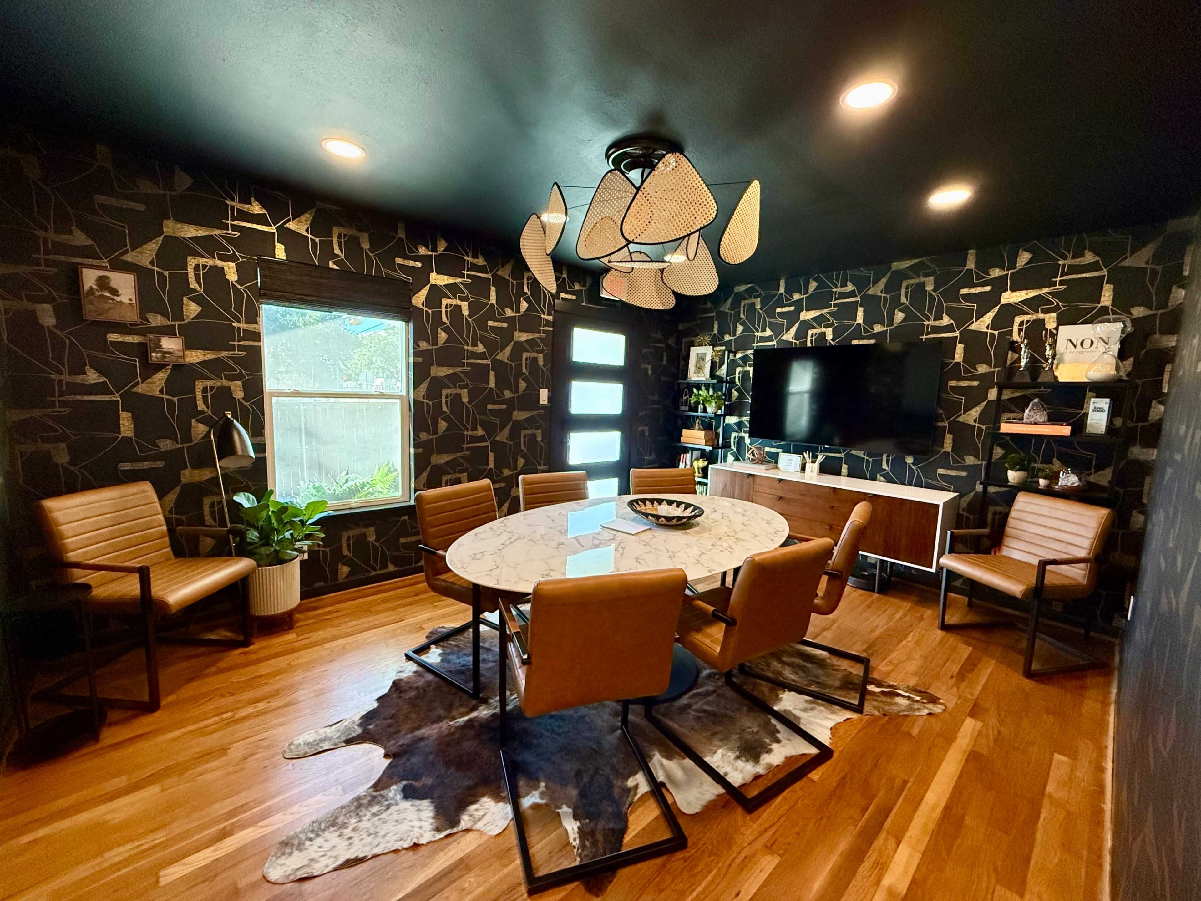 The room features a circular table surrounded by six leather chairs, with a patterned black wallpaper, wooden flooring, and a variety of decor including a television and plant.
