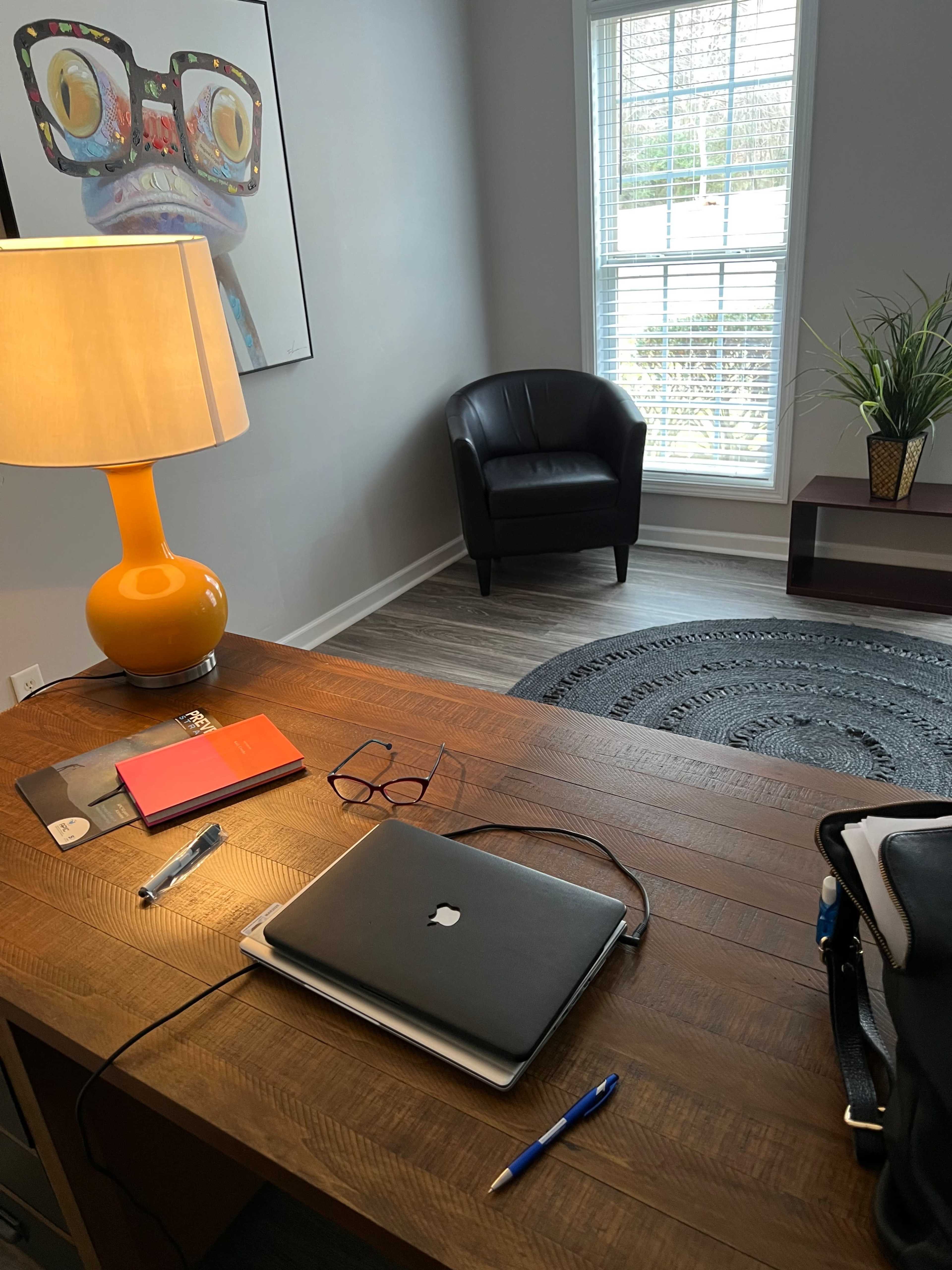 A wooden desk with a laptop, notebooks, and pens is positioned in a well-lit room featuring a chair, a lamp, and a wall art piece.