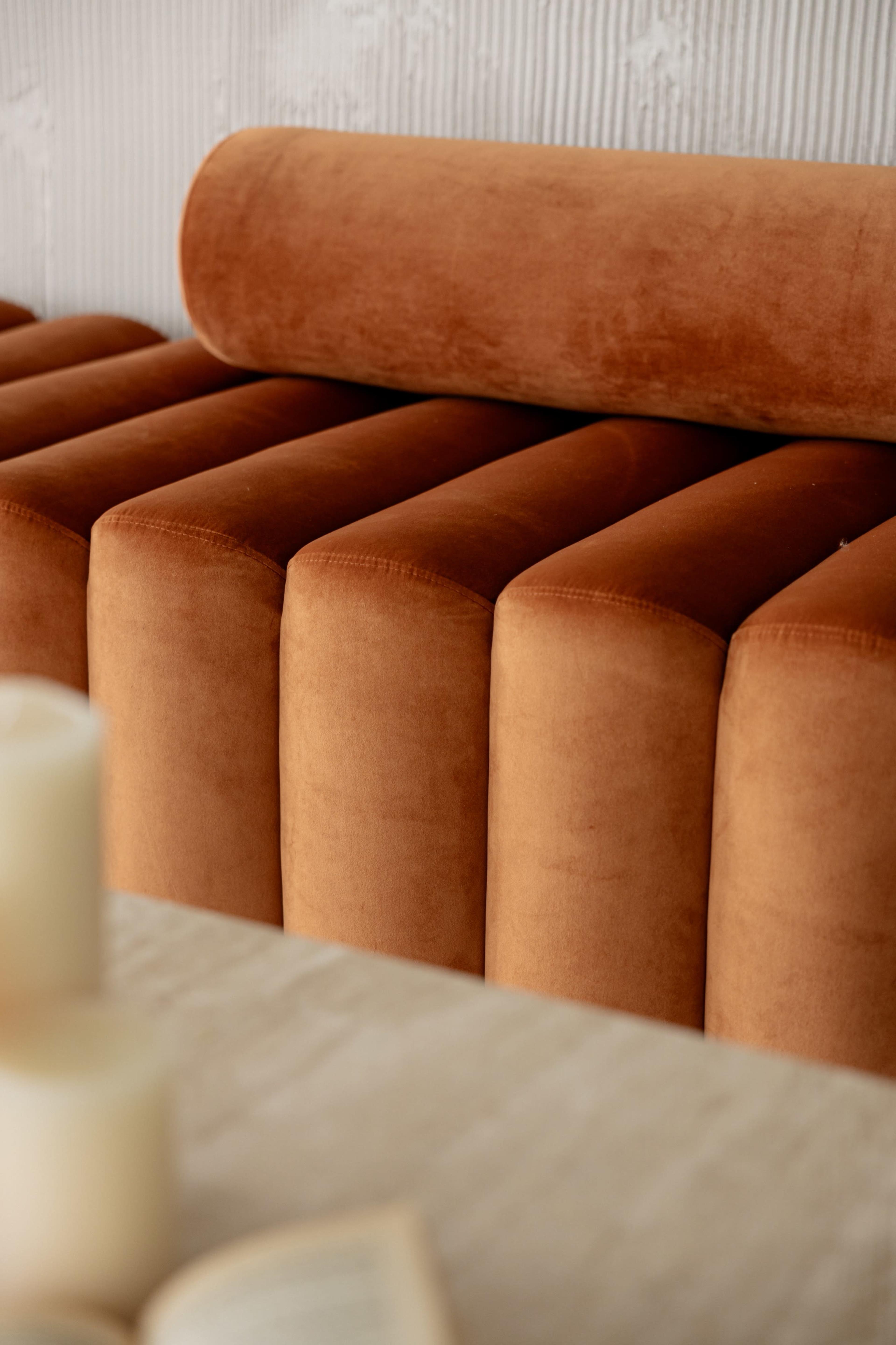 The image features a close-up of an orange velvet sofa with a cylindrical backrest and a smooth surface, placed against a textured wall.