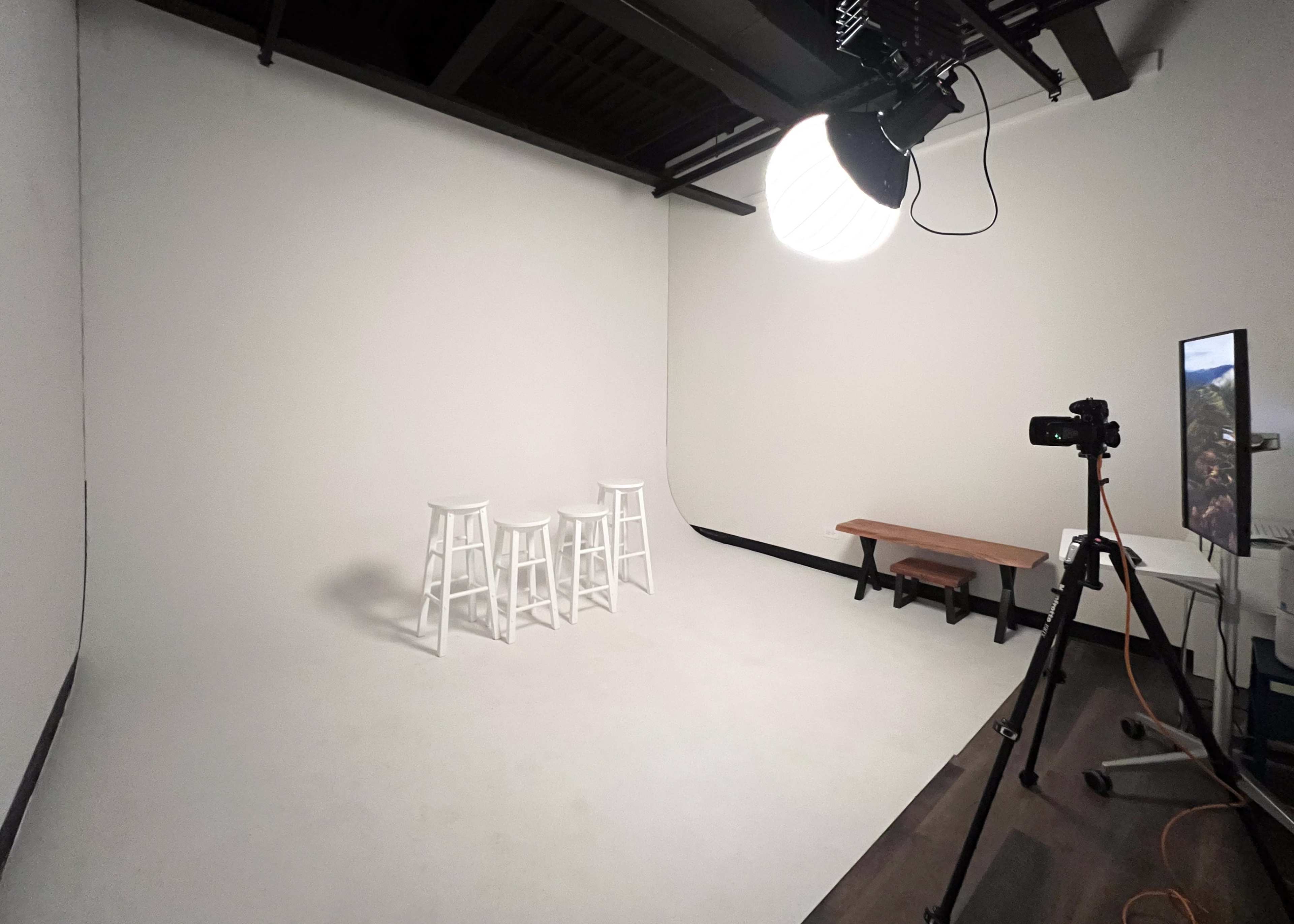 The image shows a minimalist studio space featuring three white stools, a wooden bench, and a camera setup in front of a plain white backdrop.