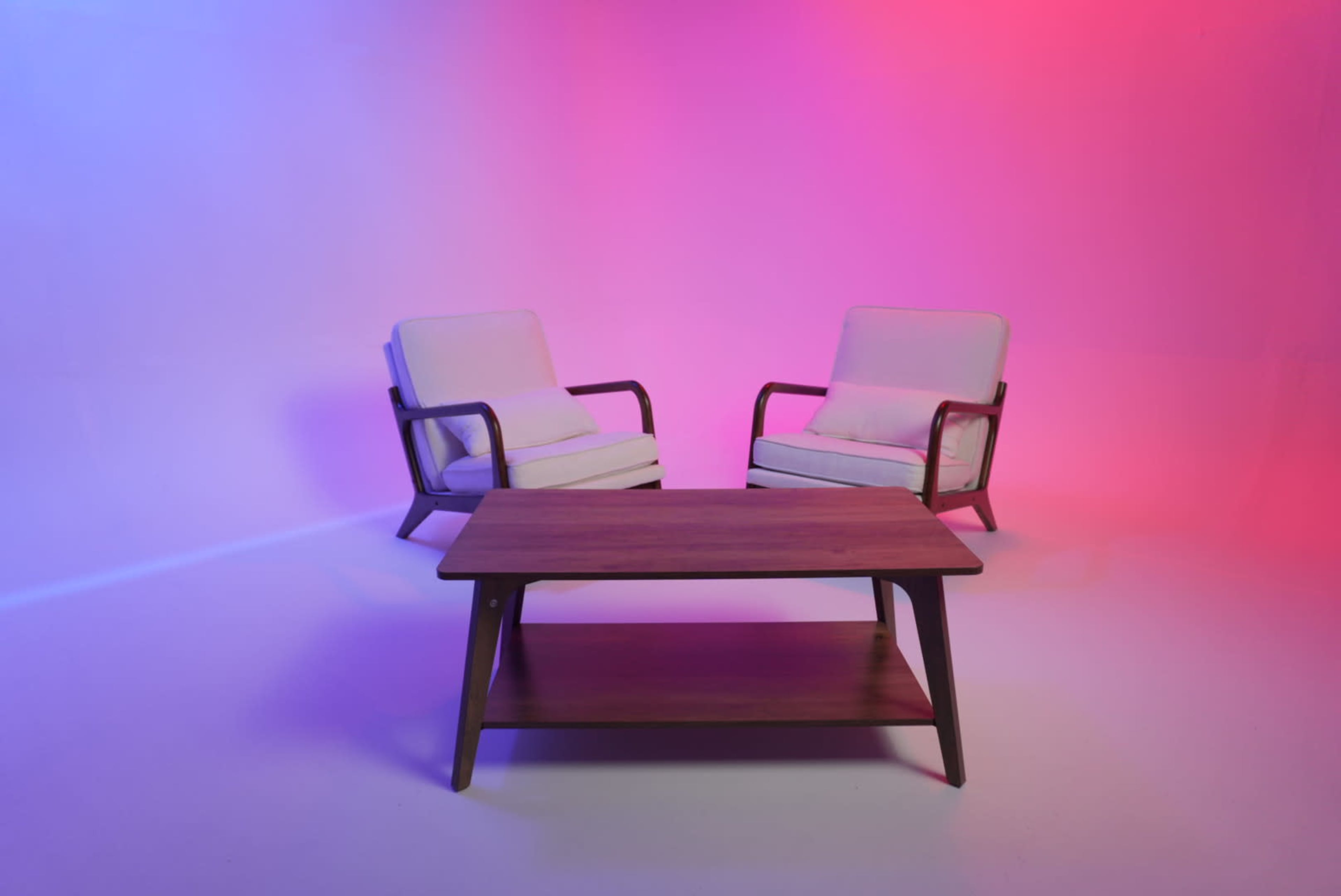 Two white chairs with wooden frames are positioned on either side of a wooden coffee table against a gradient background of purple and pink lighting.