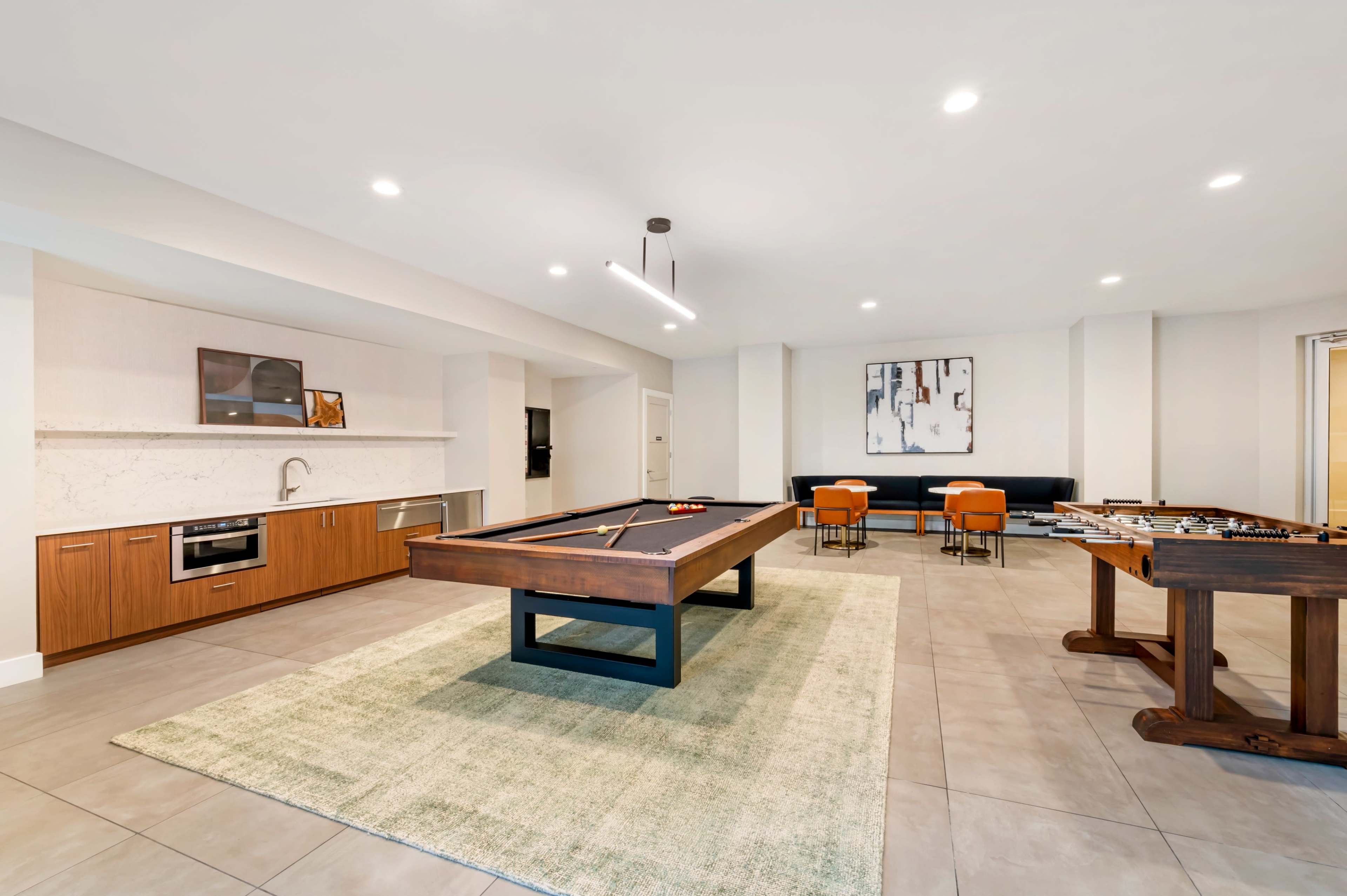 The image depicts a spacious game room featuring a billiards table, a foosball table, and seating arrangements with modern decor.