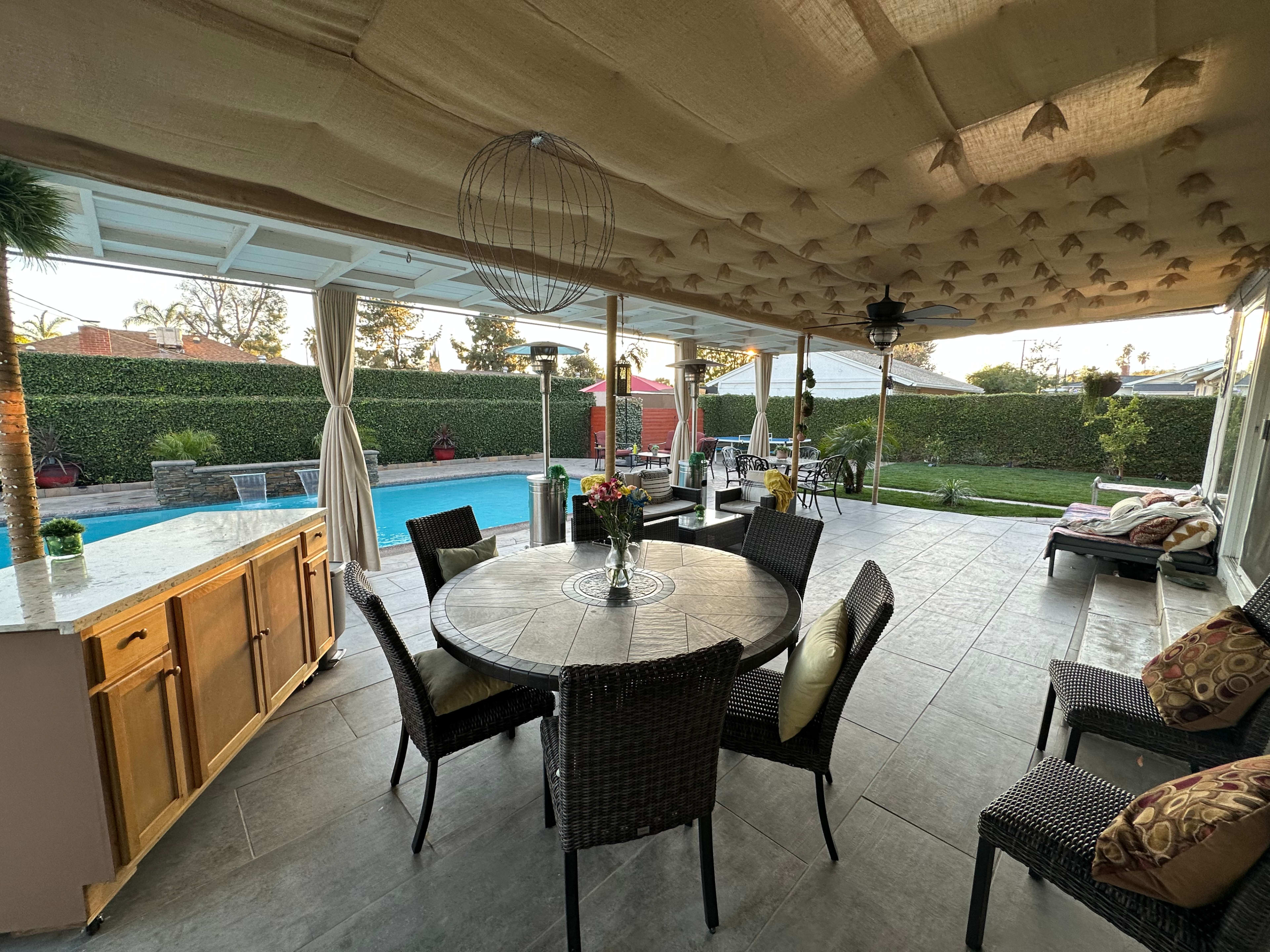 A covered patio area features a round dining table with wicker chairs, overlooking a backyard with a swimming pool and neatly trimmed hedges.
