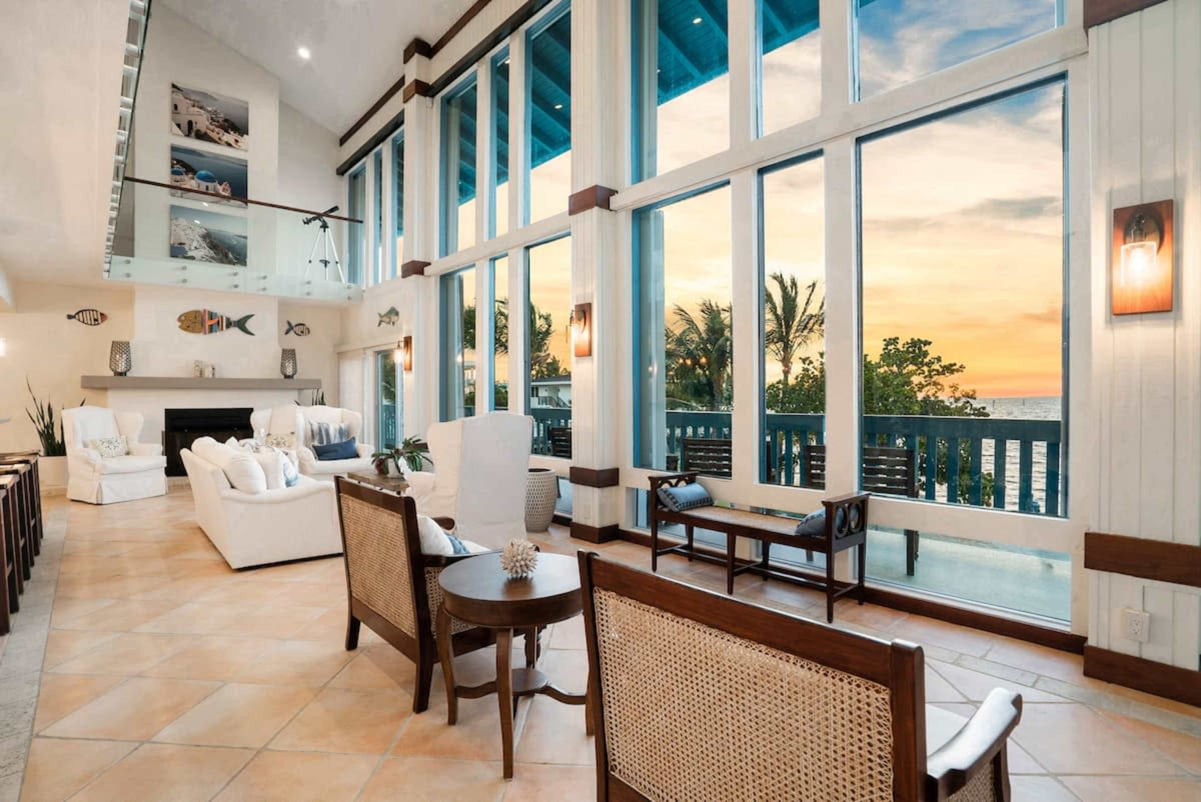 The image shows a spacious living room with large windows facing a sunset view, featuring white furniture and light-colored tile flooring.