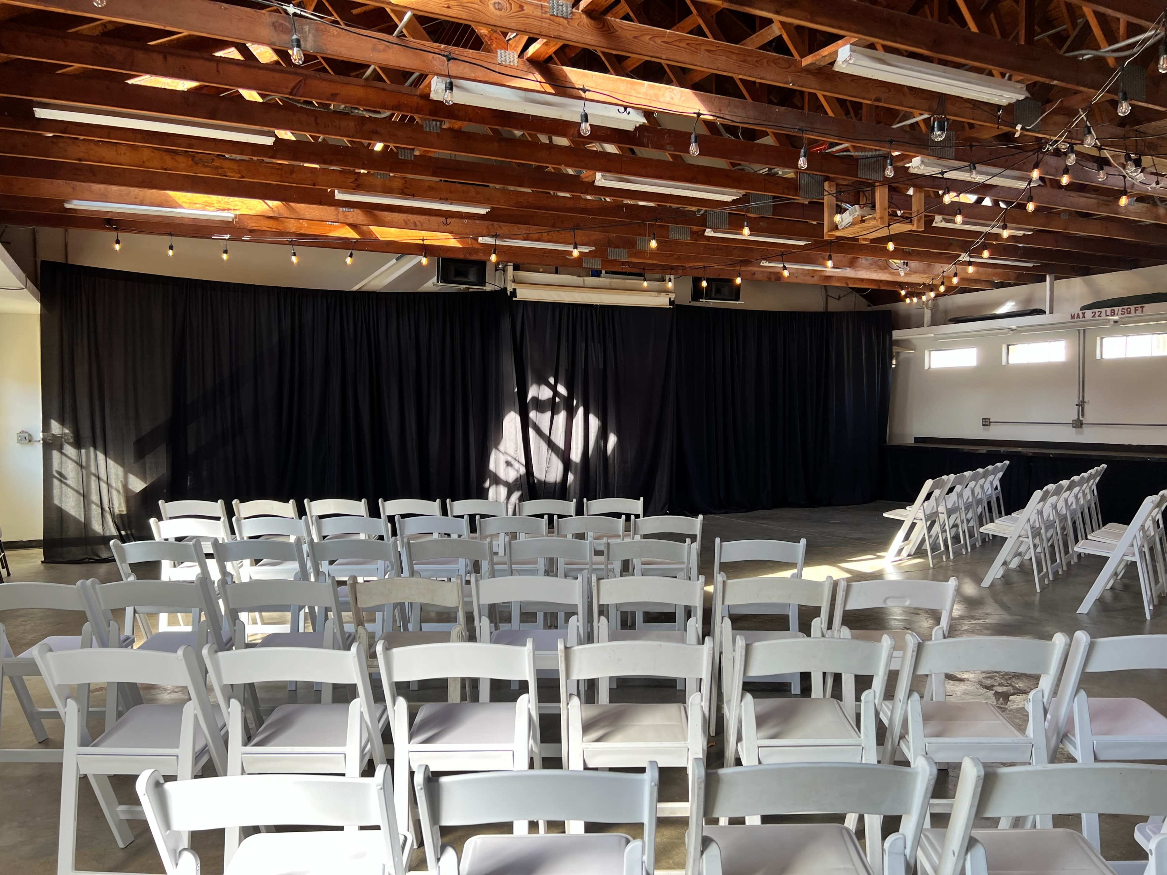 A spacious room with wooden beams and rows of white chairs facing a black curtain.