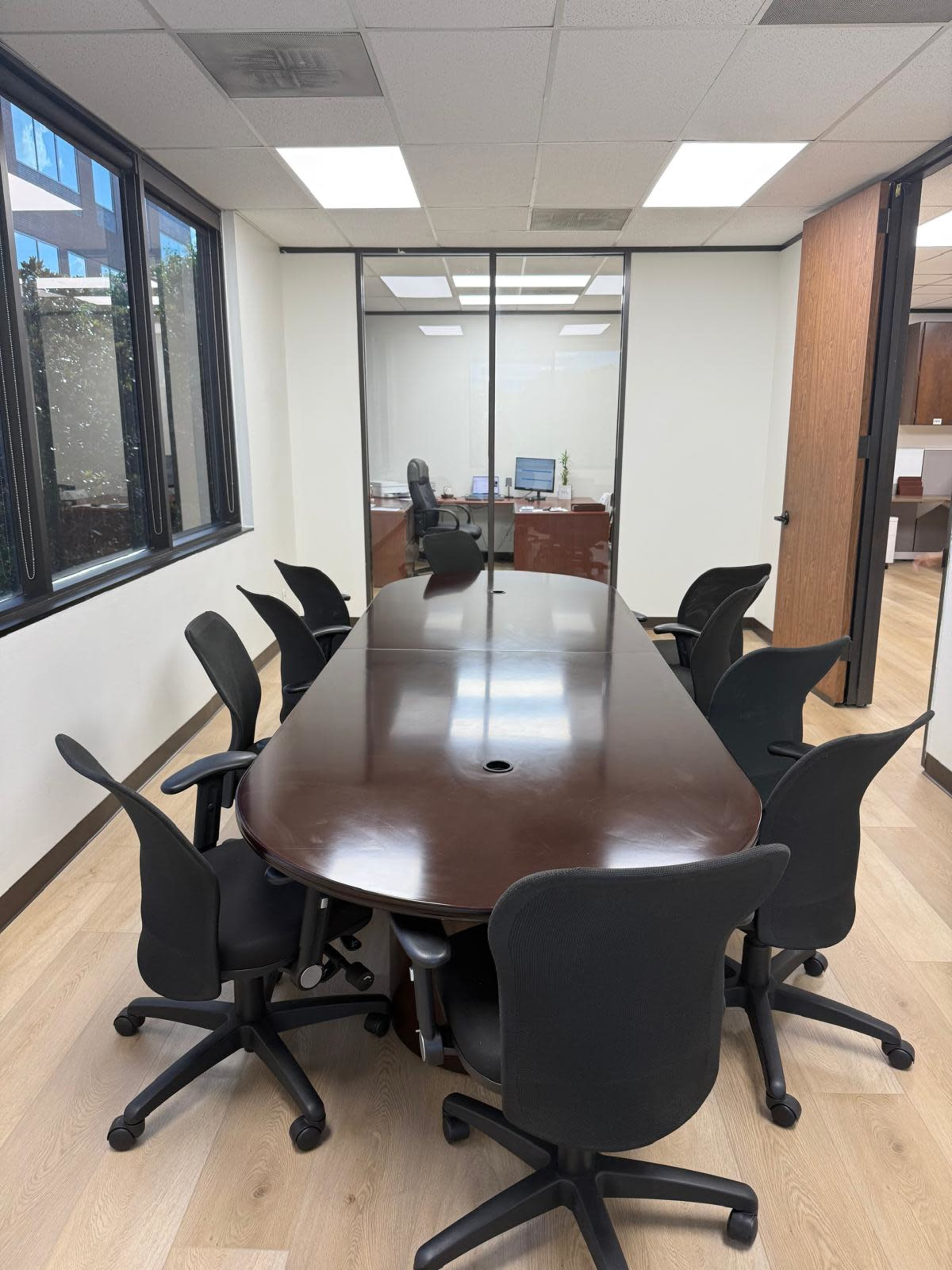 A large oval conference table surrounded by eight black rolling chairs is positioned in a well-lit meeting room with windows and an adjacent office visible in the background.