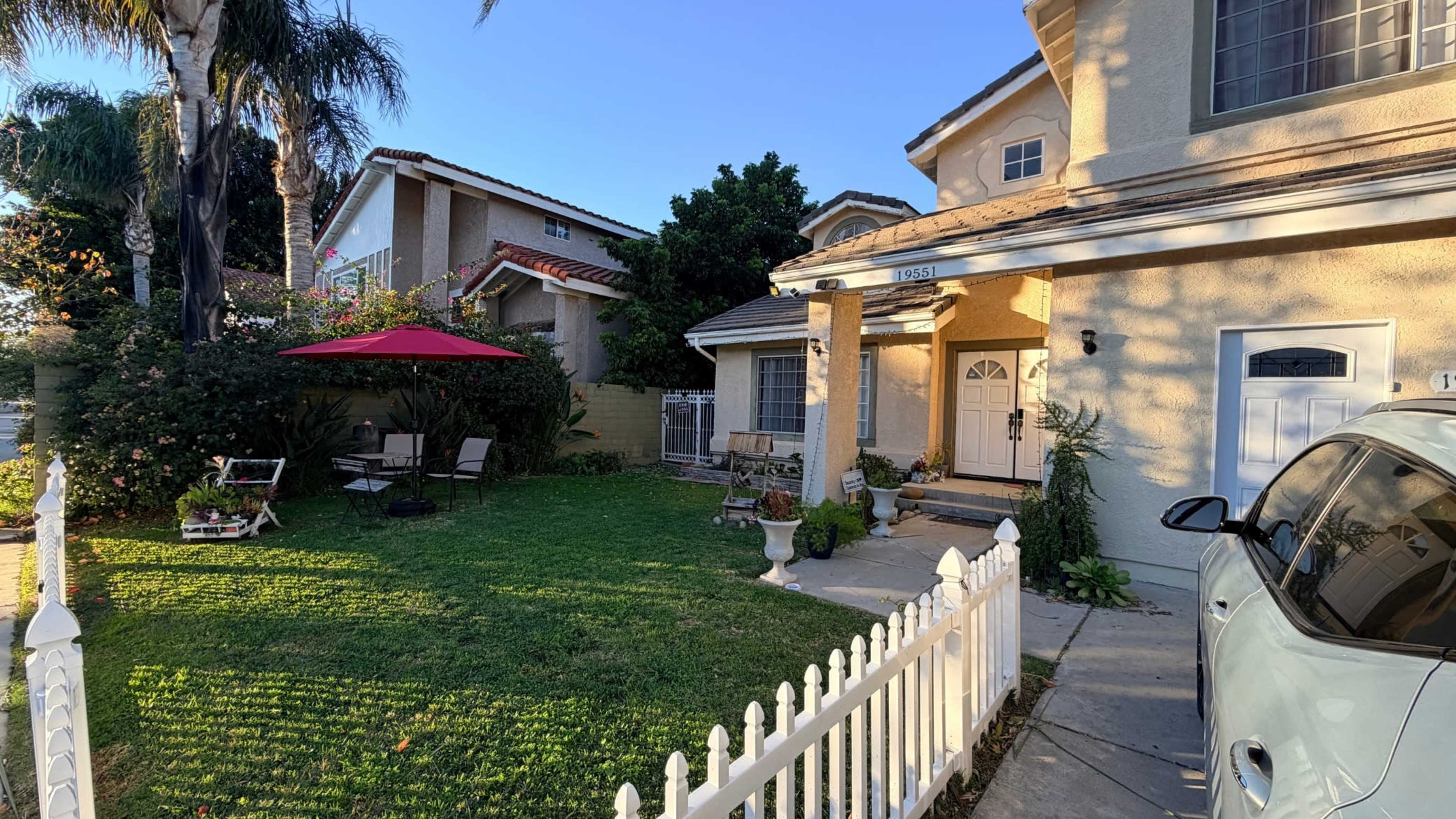 The image shows a residential property with a well-kept lawn, a white picket fence, and a red umbrella over a seating area in the front yard.