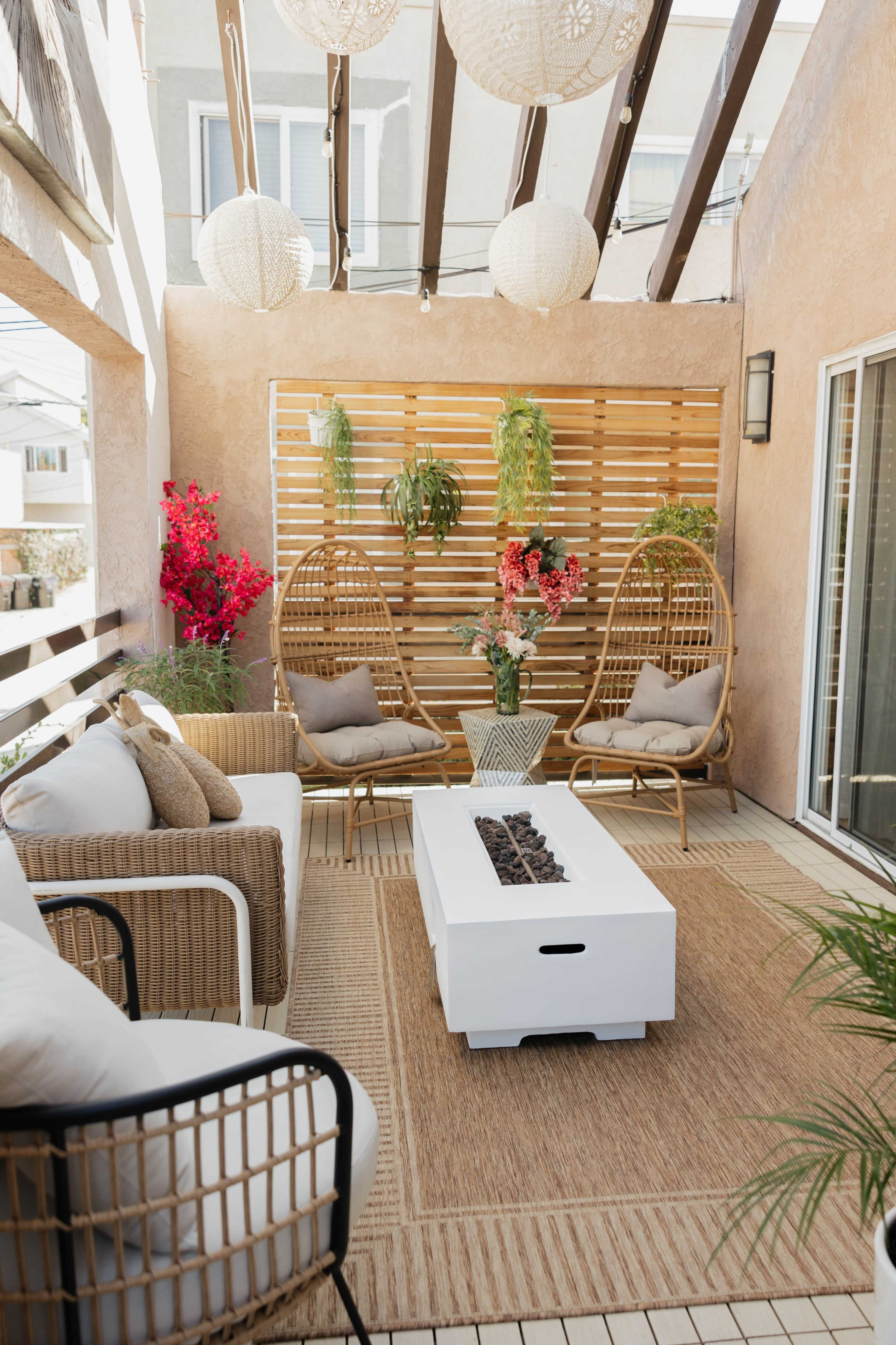 A furnished outdoor patio features two hanging chairs, a white coffee table with a fire pit, and decorative plants against a wooden slatted backdrop.