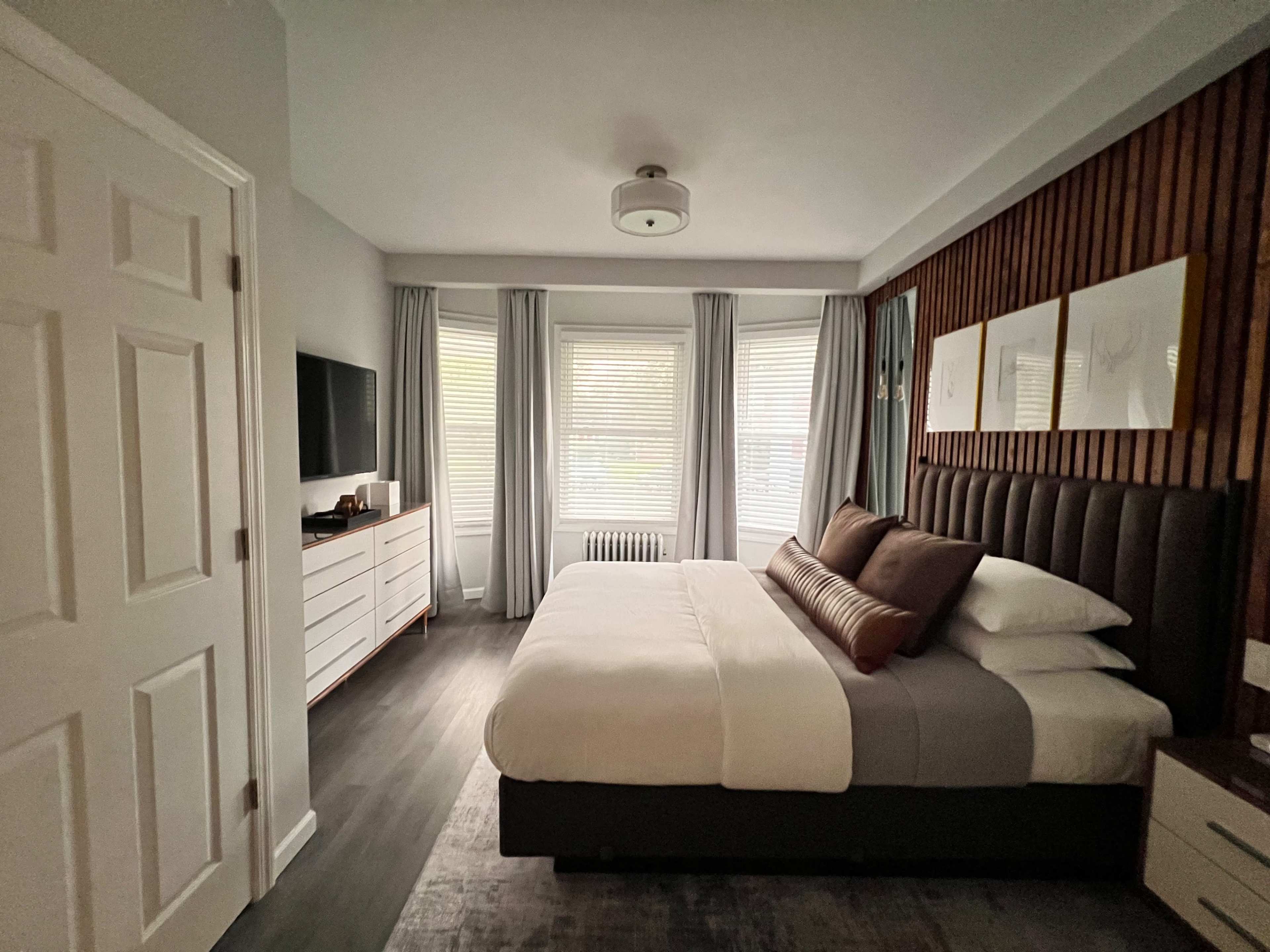 A modern bedroom features a large bed with gray and white bedding, a wall-mounted TV, and tall windows dressed with light curtains.