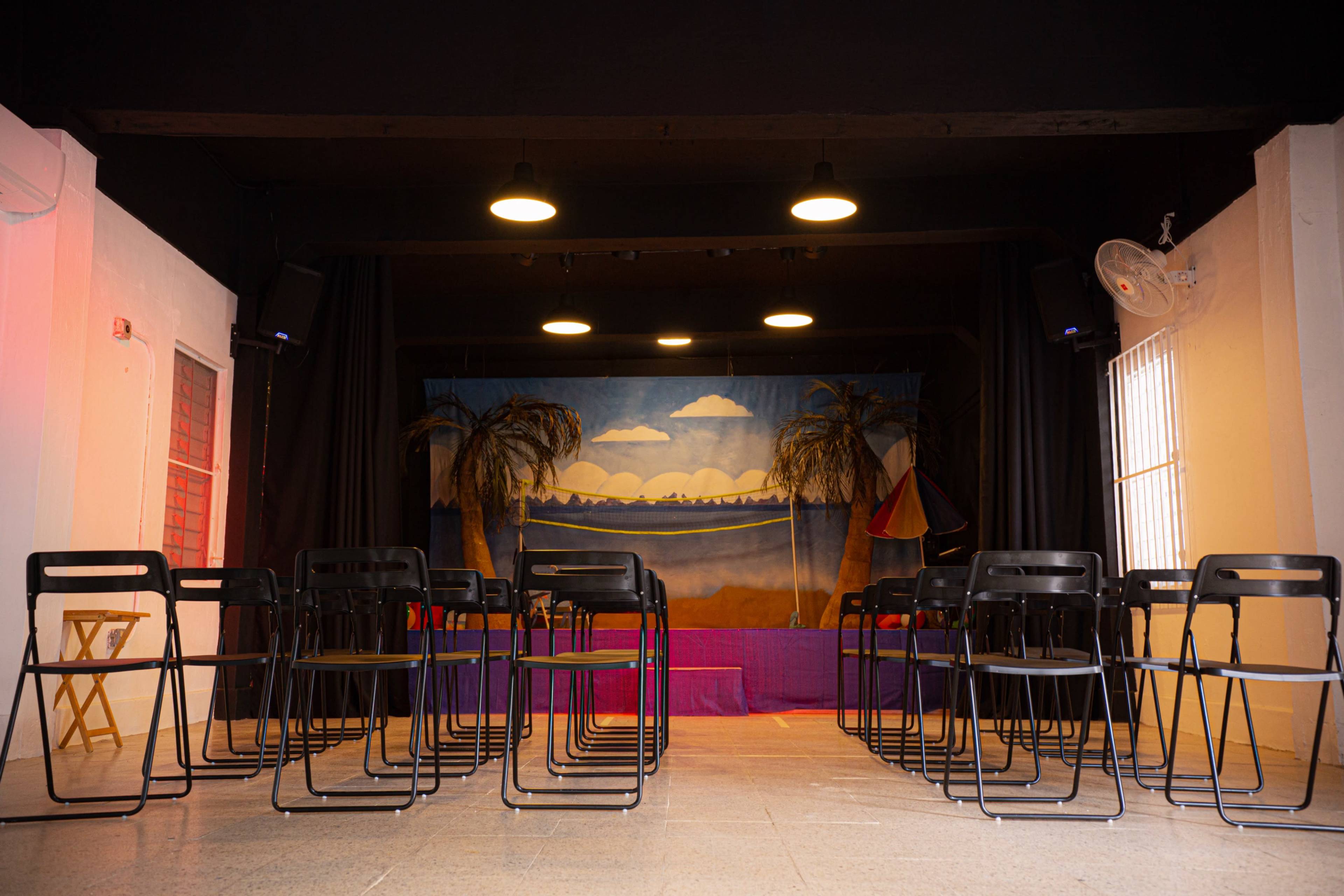 A theater stage with a backdrop of palm trees and clouds is set up in front of a row of black folding chairs arranged in the audience area.