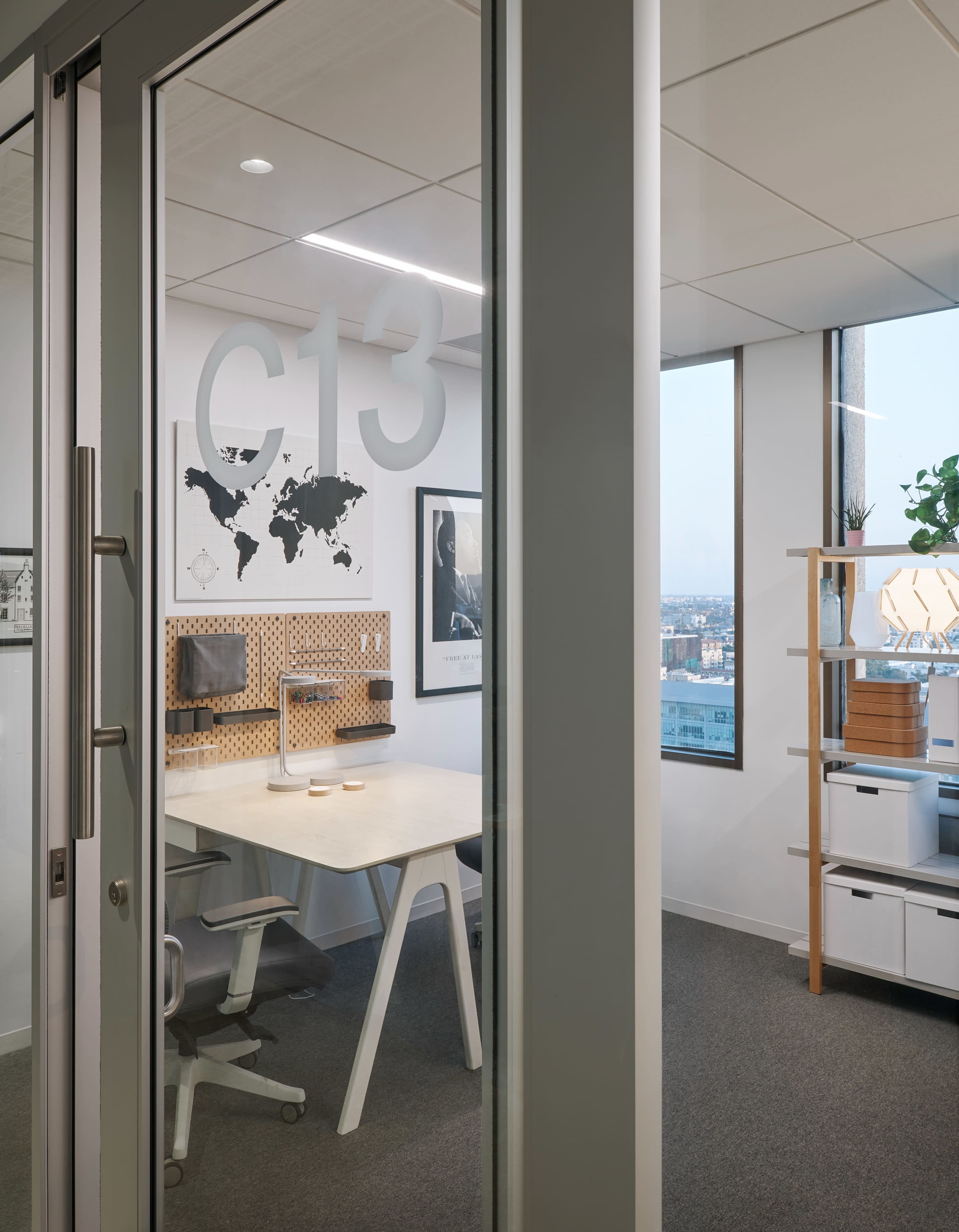 A glass-walled office with a conference table, chairs, and a map on the wall overlooks a cityscape through a large window.