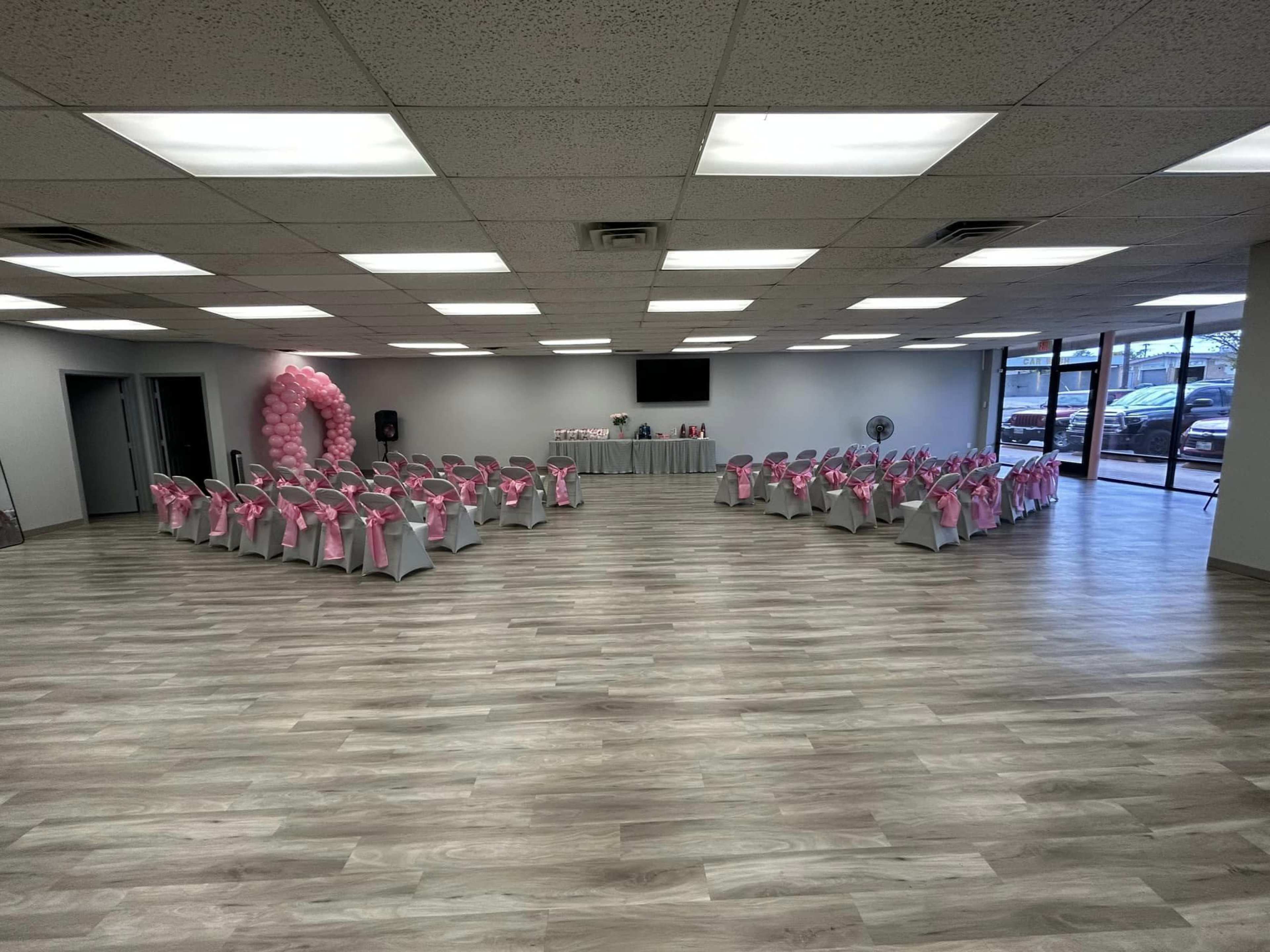 A spacious event room arranged with chairs covered in white cloth and pink sashes, set up in two rows facing a central area decorated with balloons and a table.
