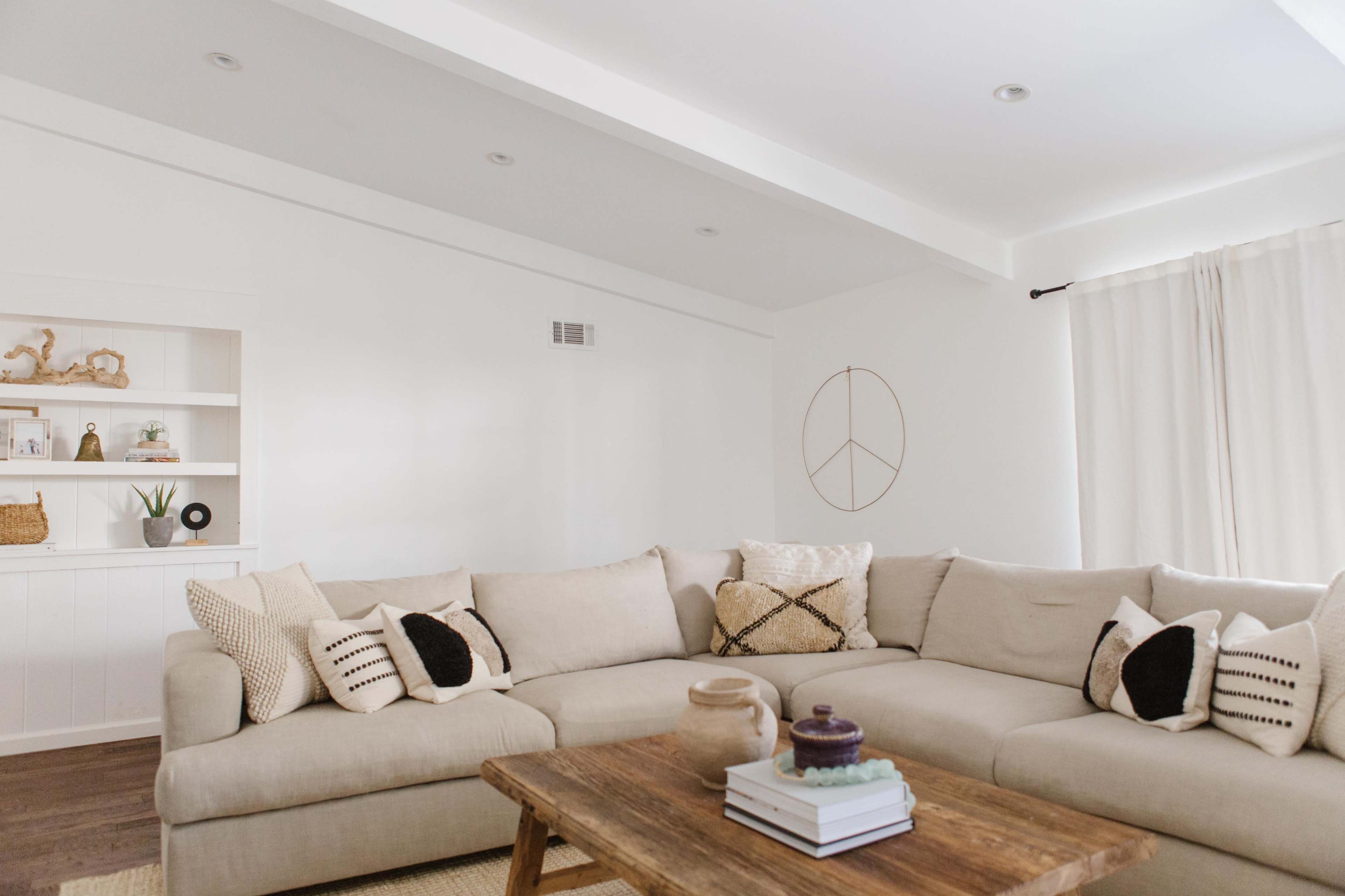 A light-colored living room featuring a large, L-shaped sectional sofa with decorative pillows, a wooden coffee table, and a shelf with a few decorative items against a white wall.