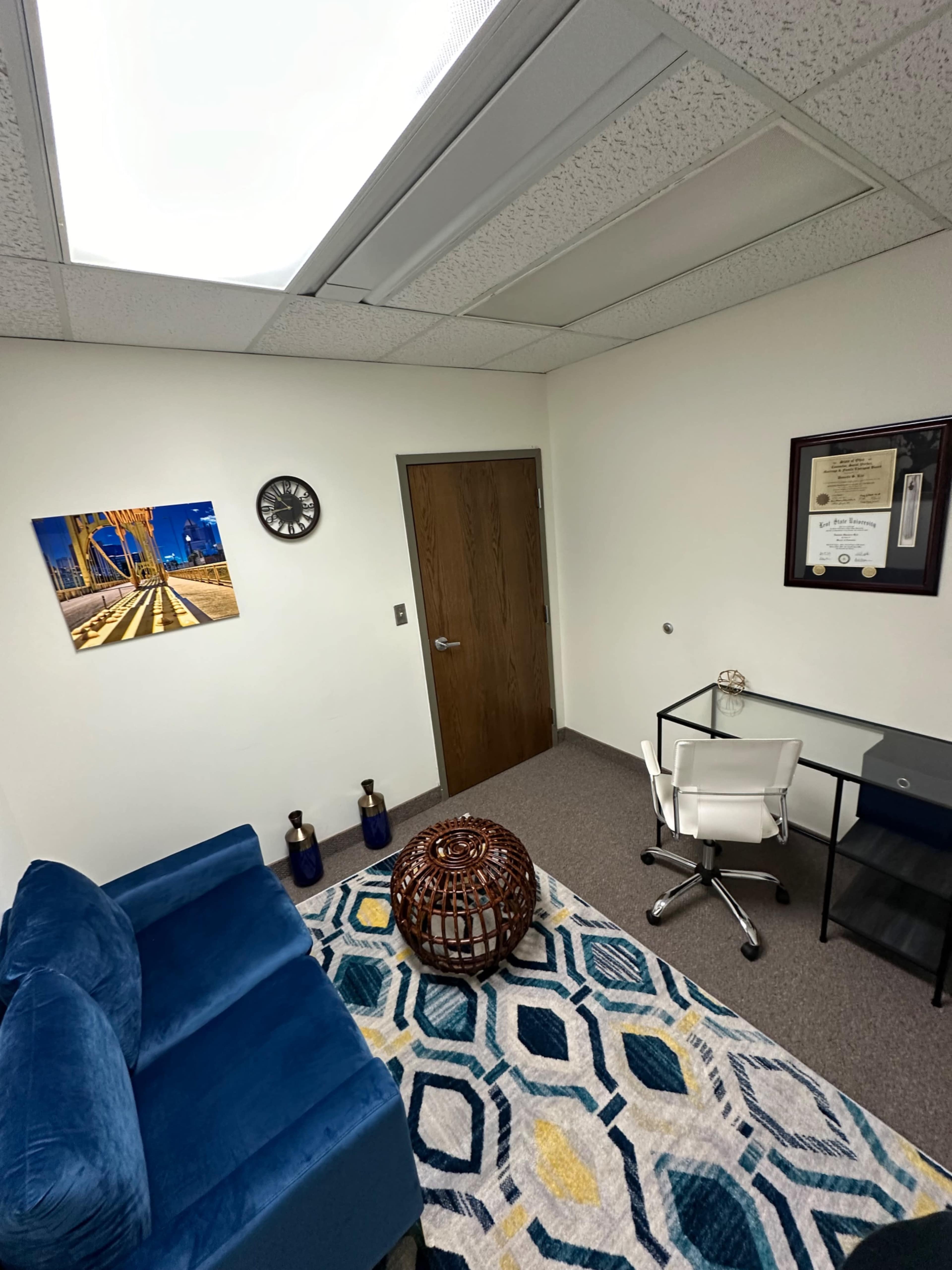 A small office featuring a blue couch, a glass desk with a chair, a patterned rug, and framed wall art.