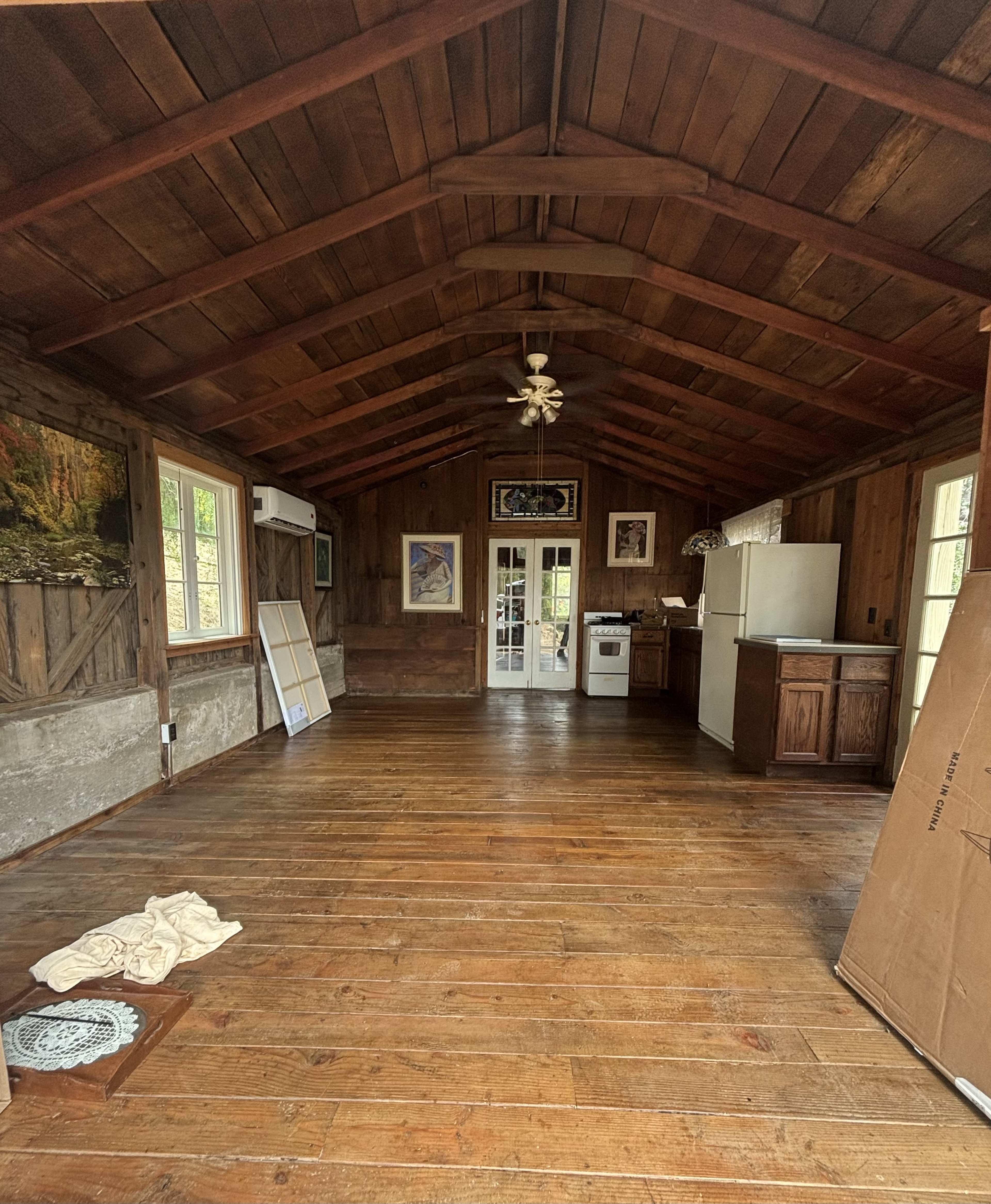 The image shows the interior of a rustic wooden cabin with a high ceiling, featuring a kitchen area and an open space with wooden flooring.