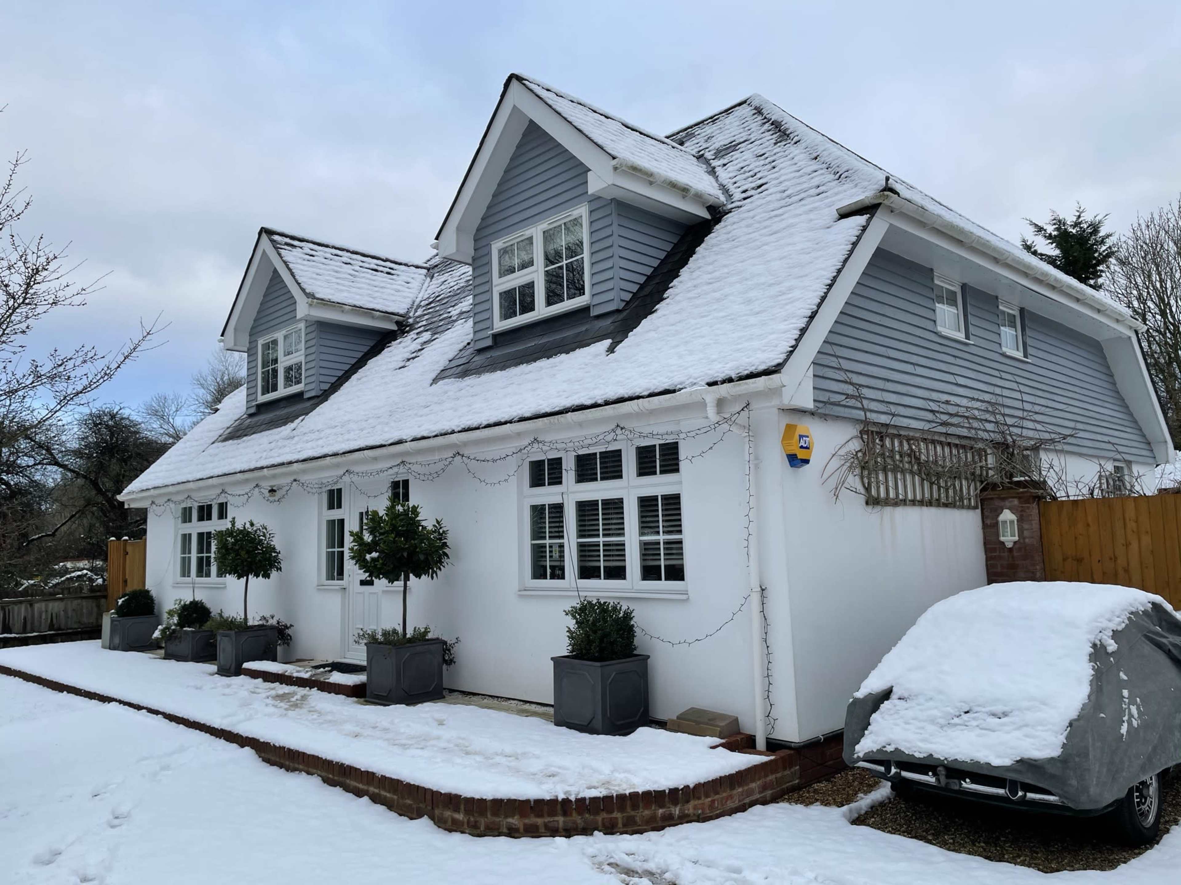 A two-story house with a grey and white exterior is surrounded by snow, featuring planters with small trees and a covered car in the driveway.