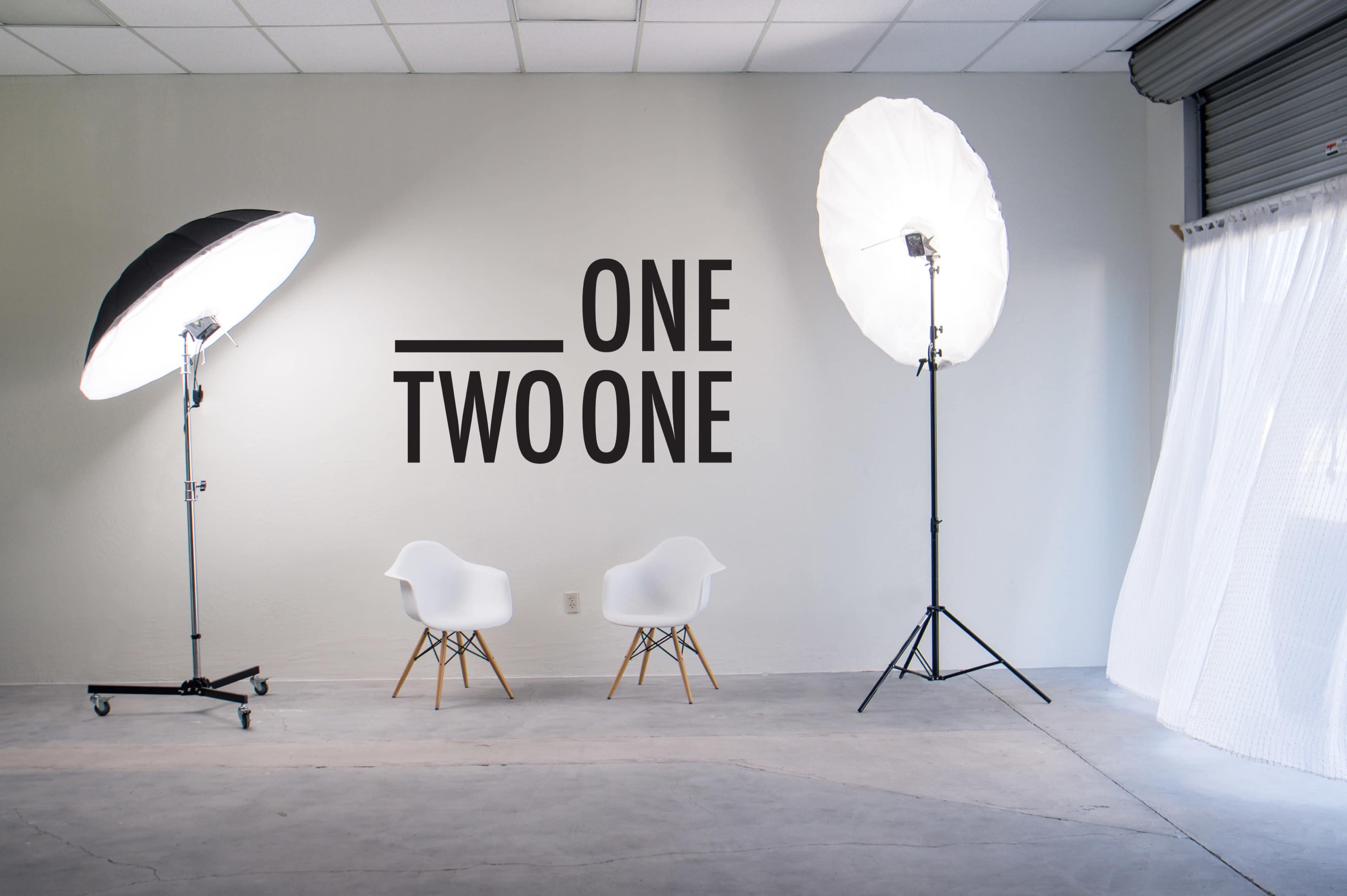 A photography studio with two softbox lights, two white chairs, and a wall featuring the text "ONE TWO ONE."