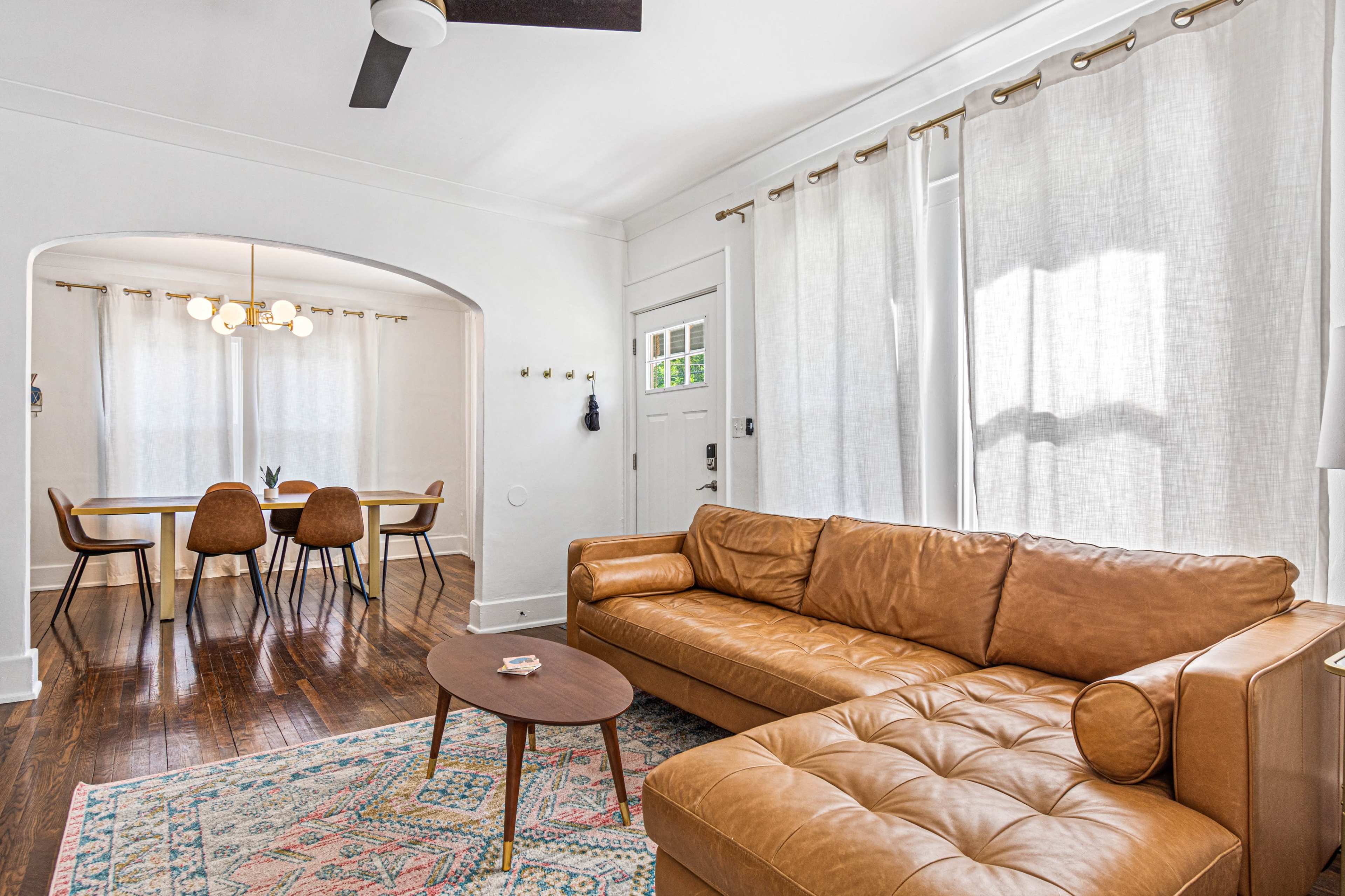 A modern living room features a brown leather sofa, a round coffee table, and light-colored curtains, with a dining area visible in the background.